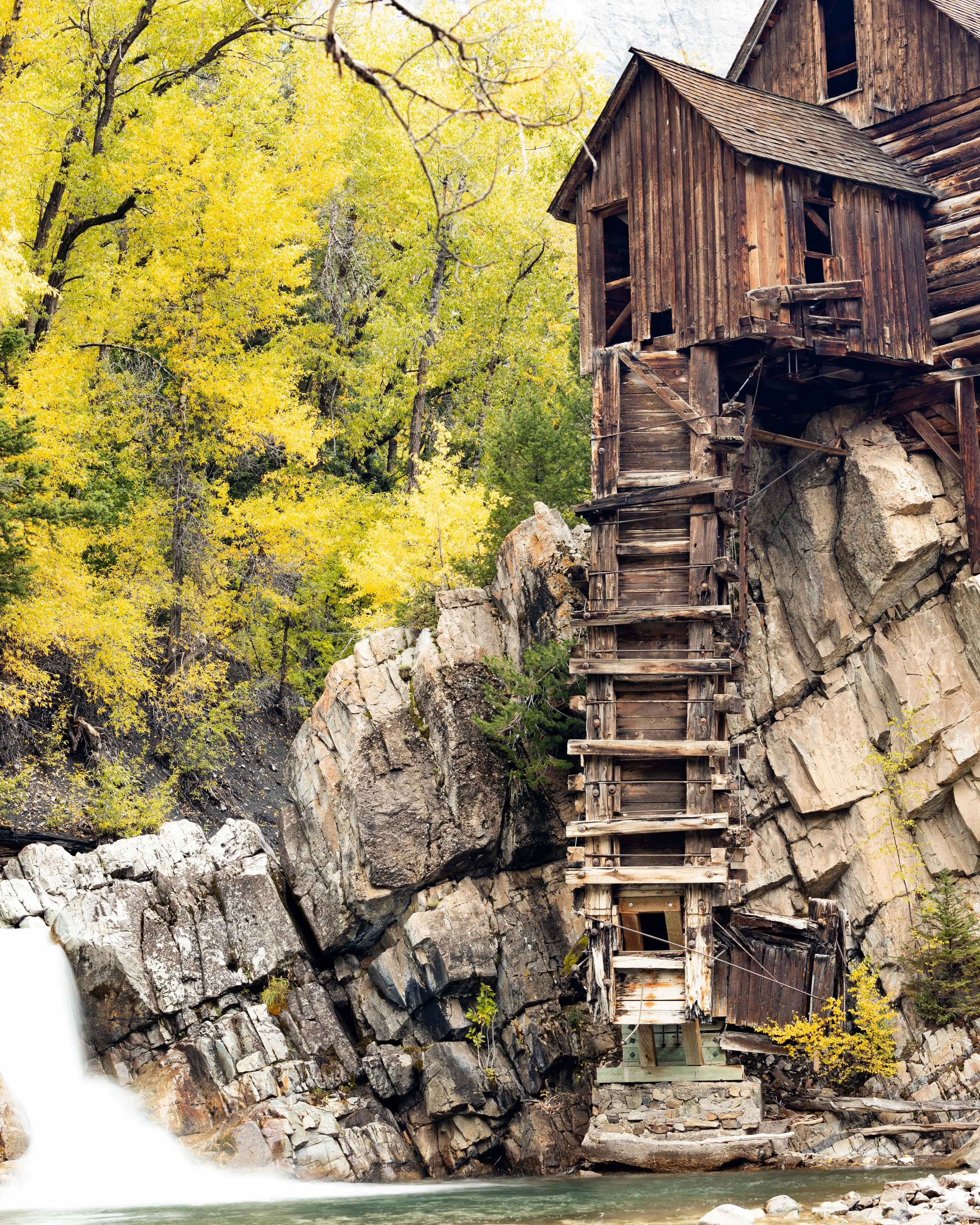 Old wooden building built on rocky cliffside near river with a small waterfall, surrounded by yellow and green autumn trees.