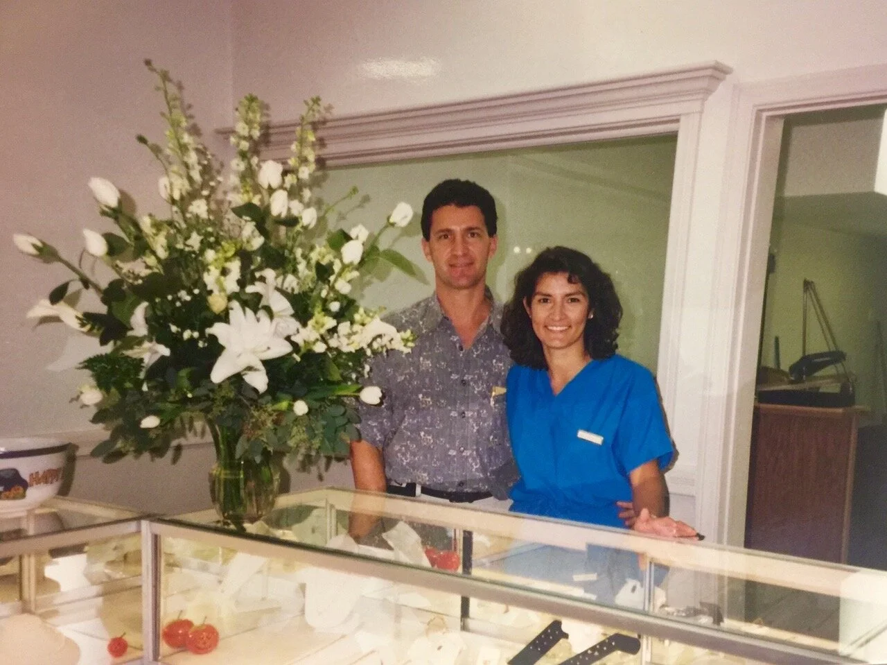 A man and a woman standing behind a glass display case with a large bouquet of white flowers beside them, in an indoor setting.