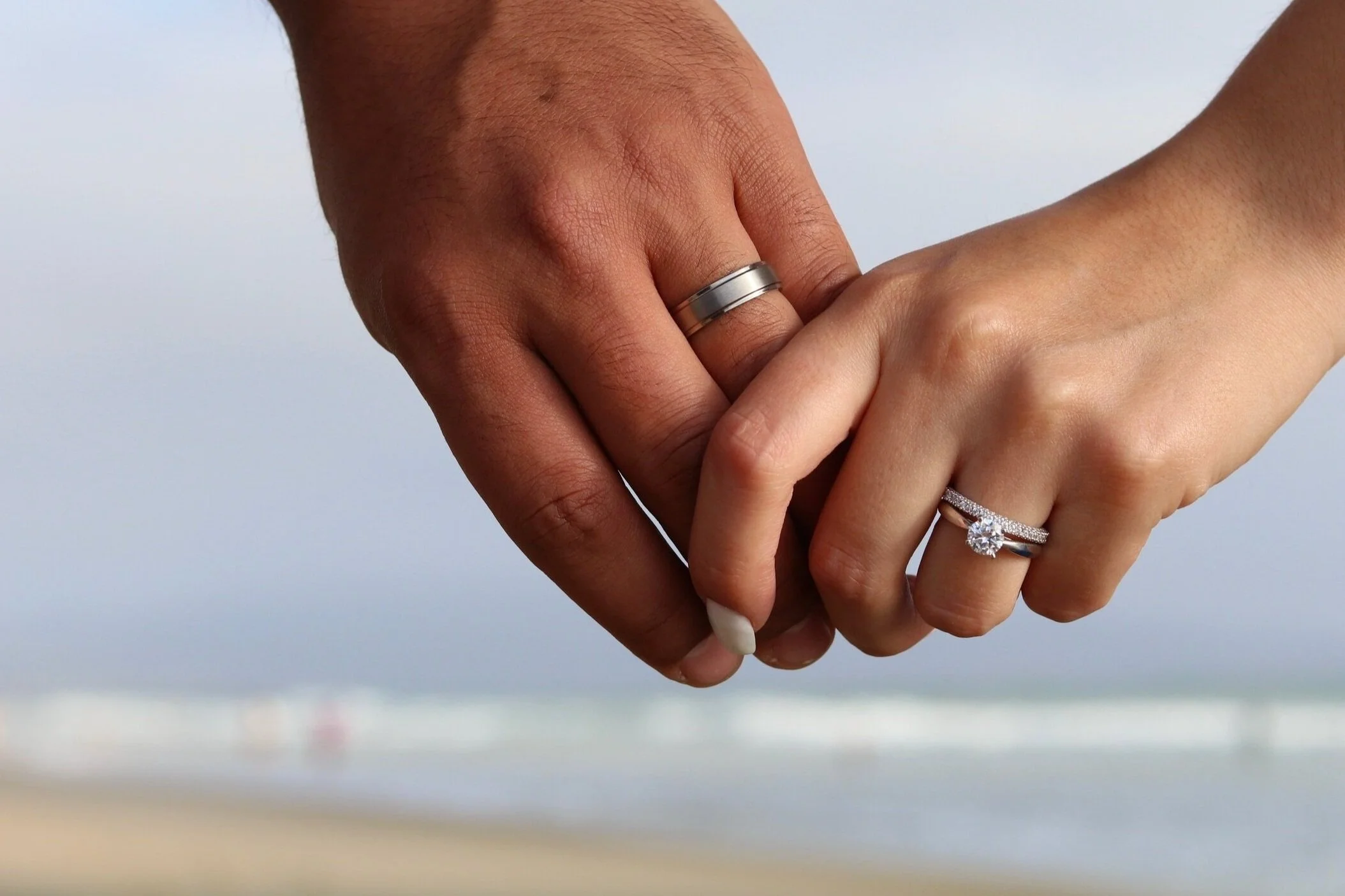 Close-up of two hands holding each other, displaying engagement rings, against a beach background.