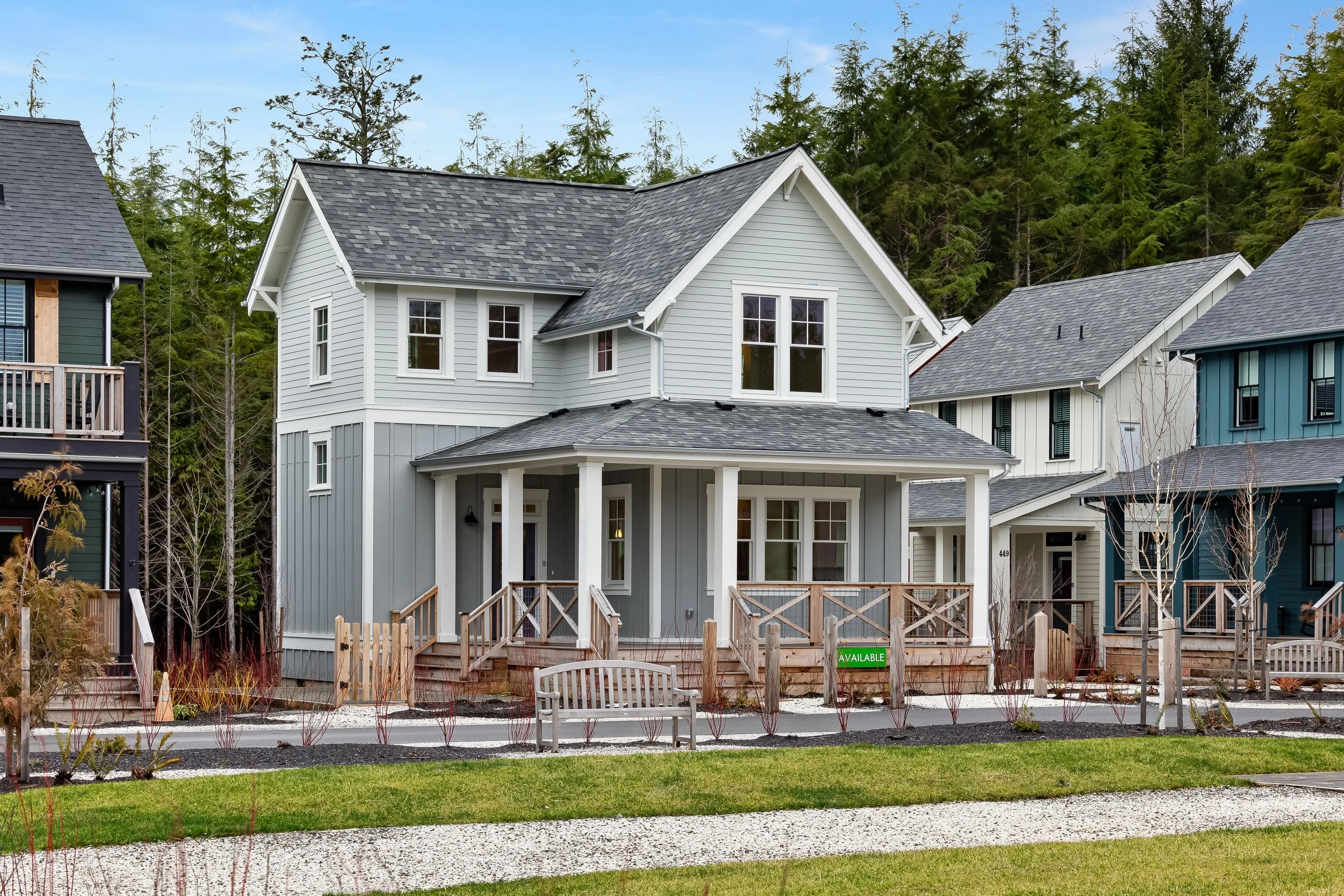 The picture of a home in Seabrook, Washington two-story white and grey house with a light gray roof sour refounded by a gravel, a green park and other high-end homes with lap siding on other side, surrounded by Evergreen trees, a large porch as well