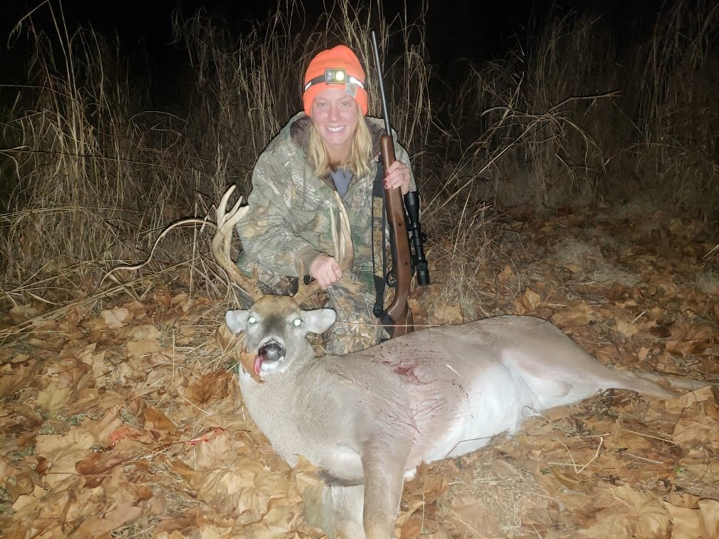 A person in camouflage and an orange hat kneeling beside a dead buck with antlers on the ground, holding a rifle, in a field with dry grasses and leaves at night.