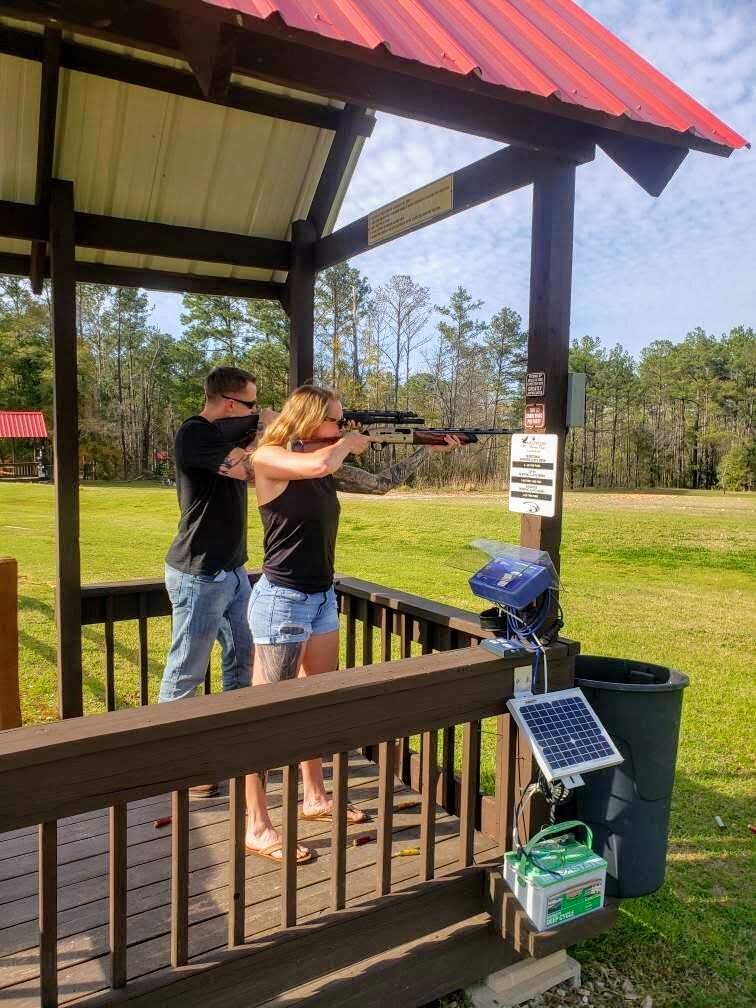Two people at a shooting range aiming rifles, standing on a wooden platform with a solar panel attached and trees in the background.