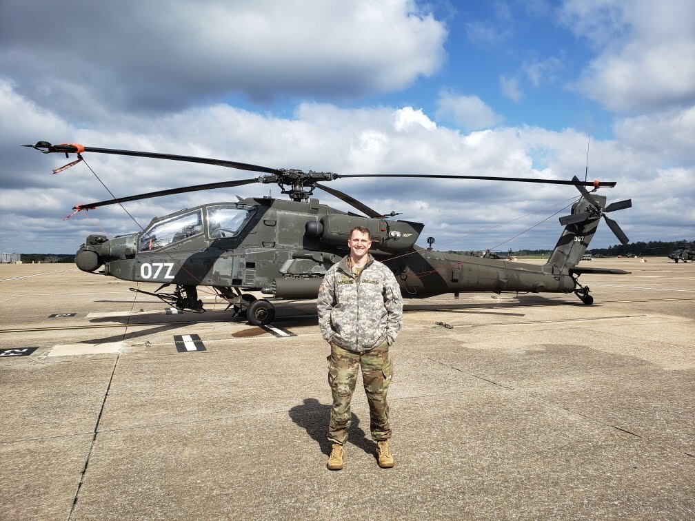 A military person standing in front of an Apache attack helicopter on a tarmac.