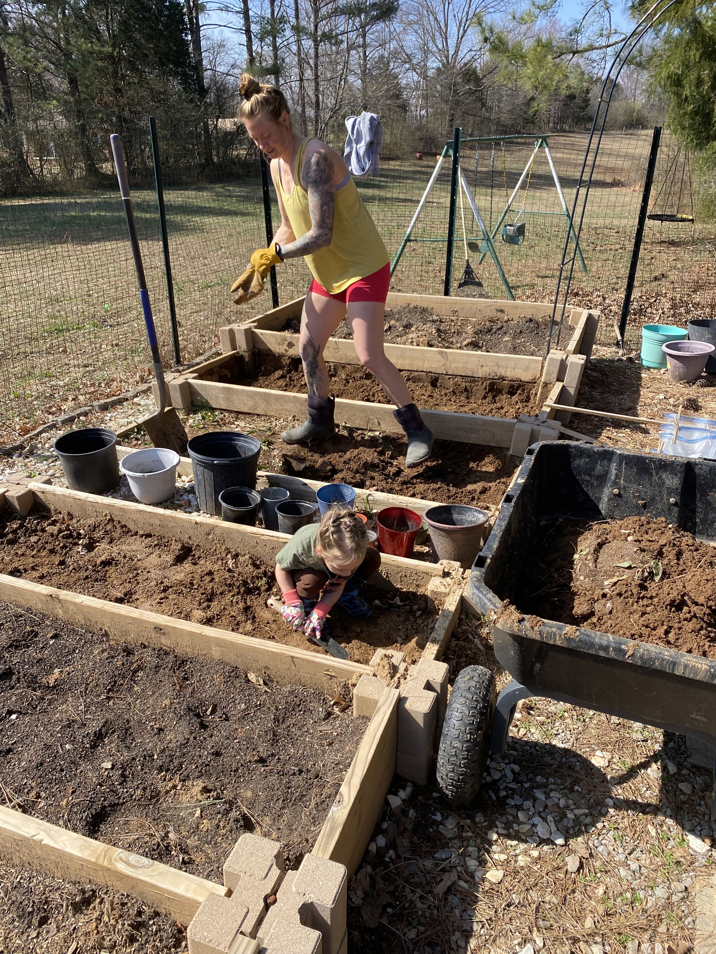 Woman in a yellow tank top and red shorts gardening with a small child in a raised bed garden, surrounded by pots and a wheelbarrow filled with soil. The garden is enclosed by a fence, and there's a swing set in the background.