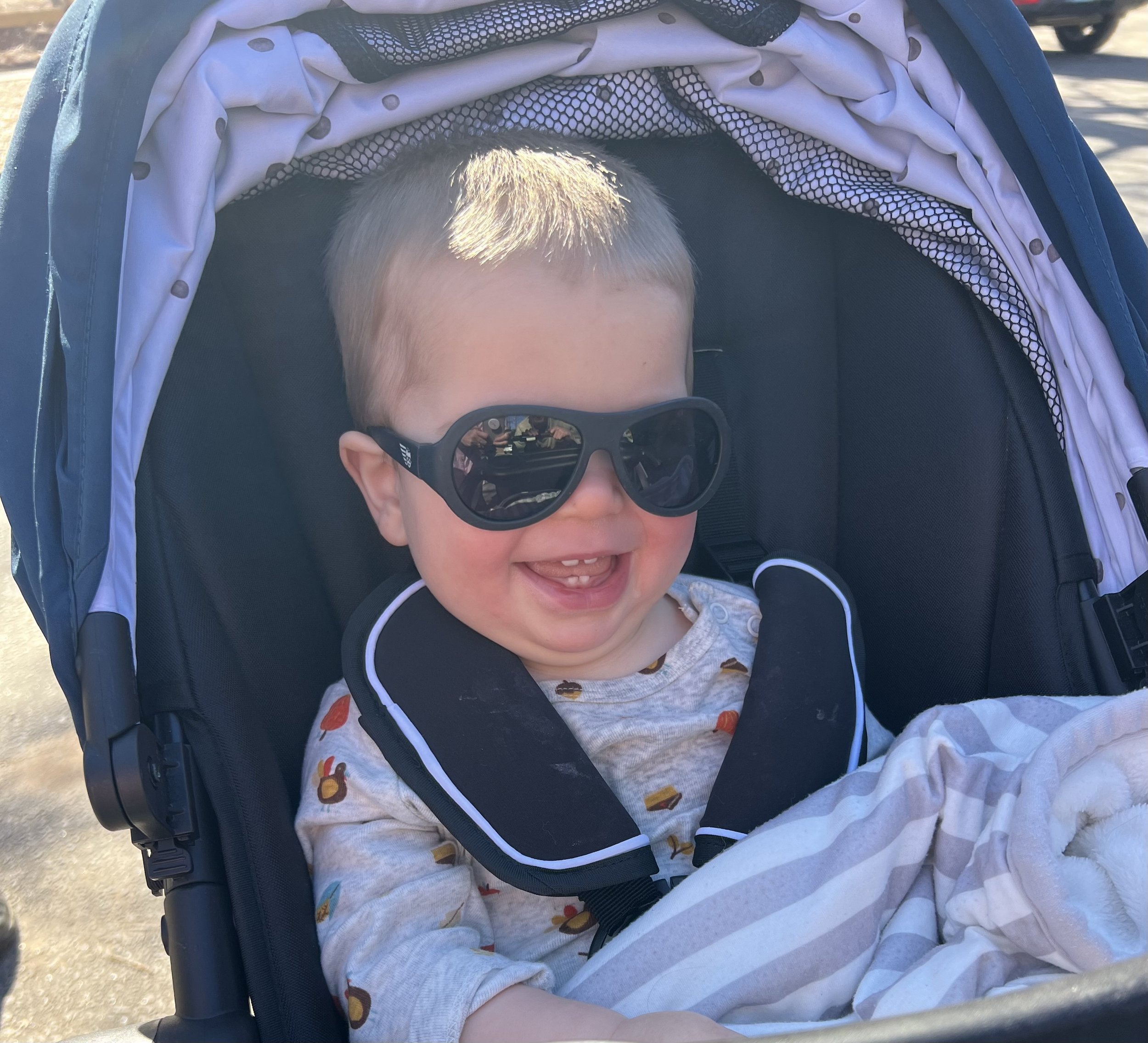 A smiling baby wearing sunglasses sitting in a stroller with a blanket.