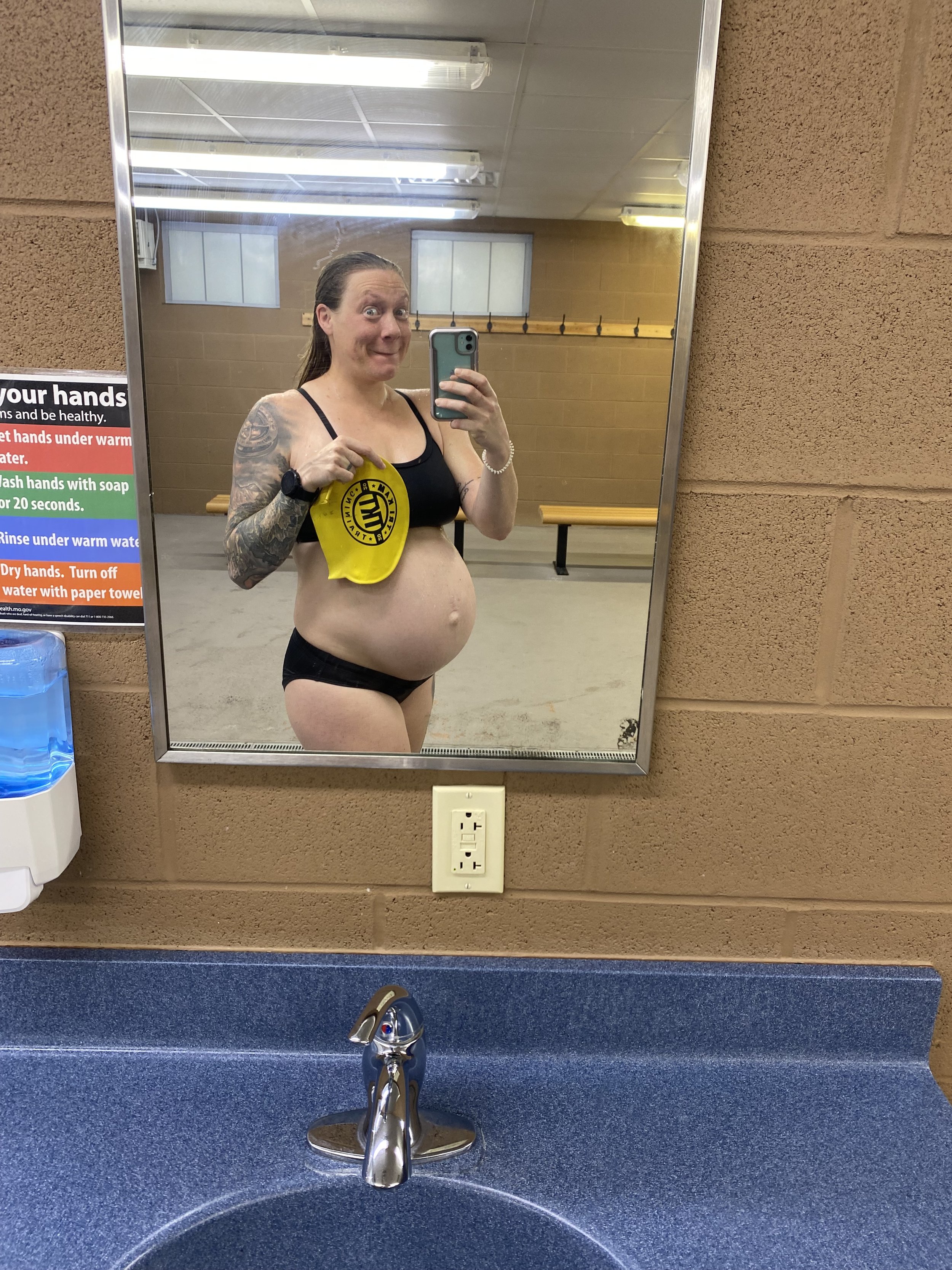 Person taking a selfie in a locker room mirror, wearing a swimsuit, holding a swim cap, with a visible tattoo on their arm.