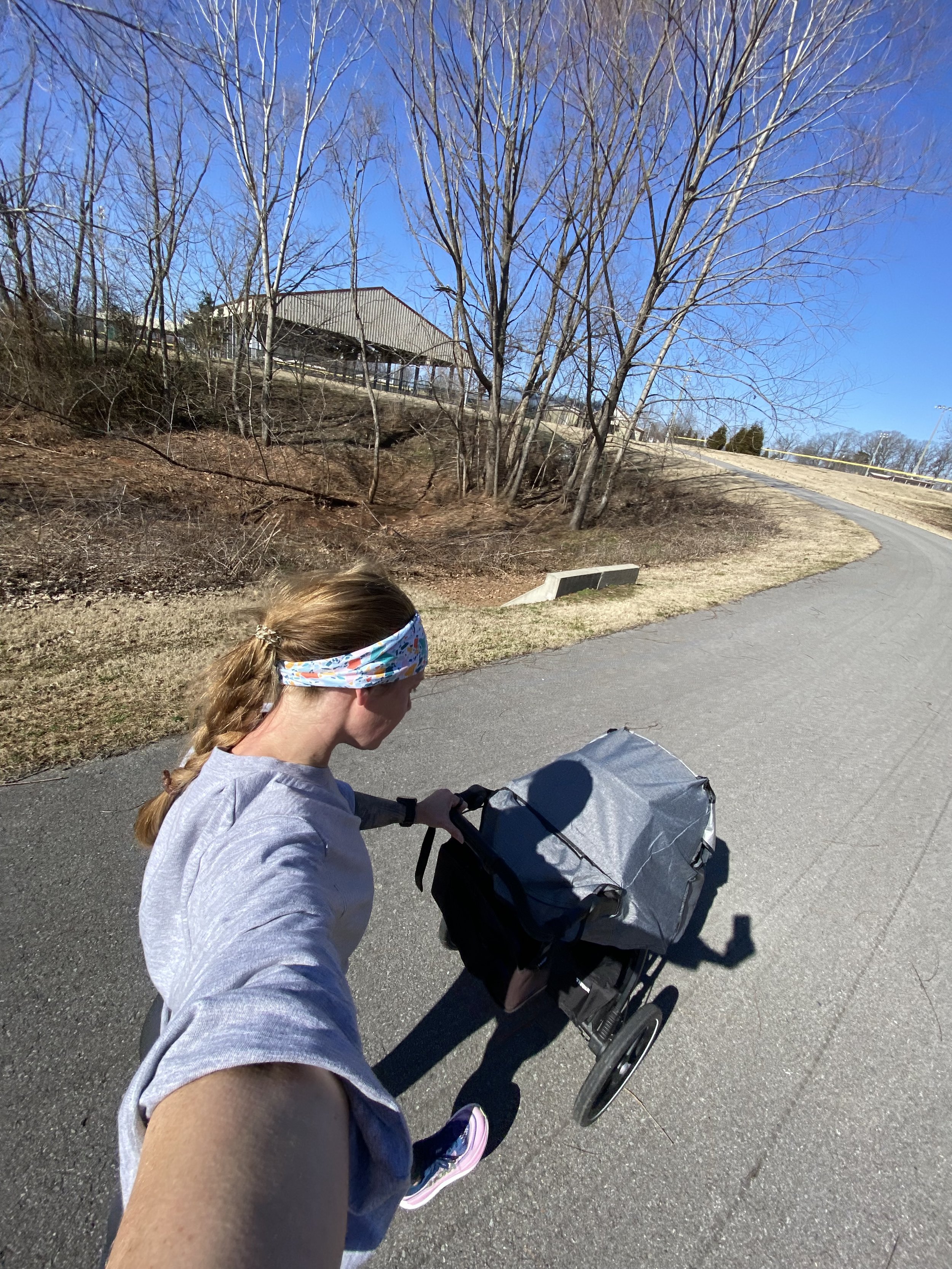 Person jogging with a stroller on a paved path, surrounded by bare trees and a pavilion in the distance on a clear, sunny day.