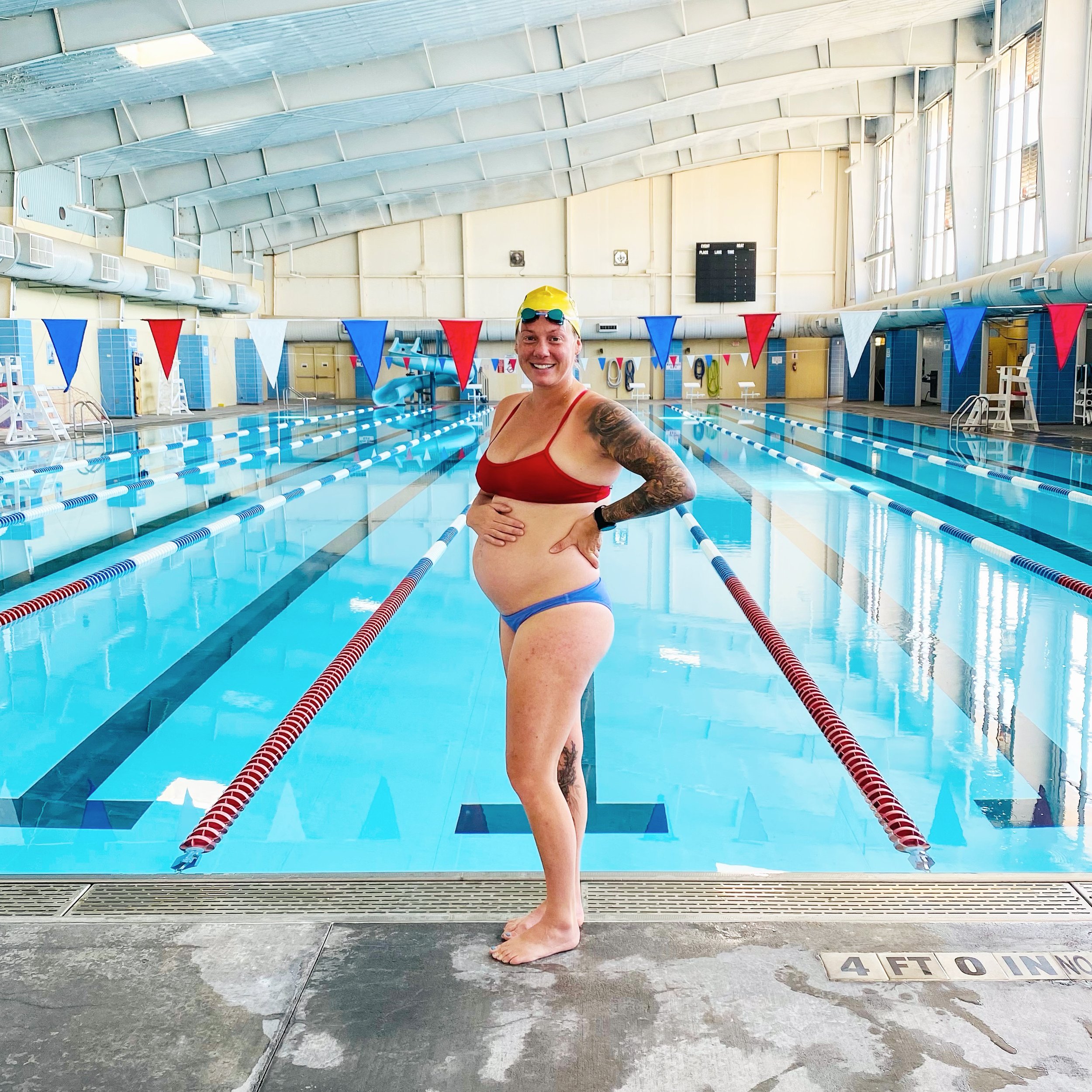 Pregnant woman in swimwear standing by an indoor pool, wearing goggles and smiling, with blue and red lane markers.