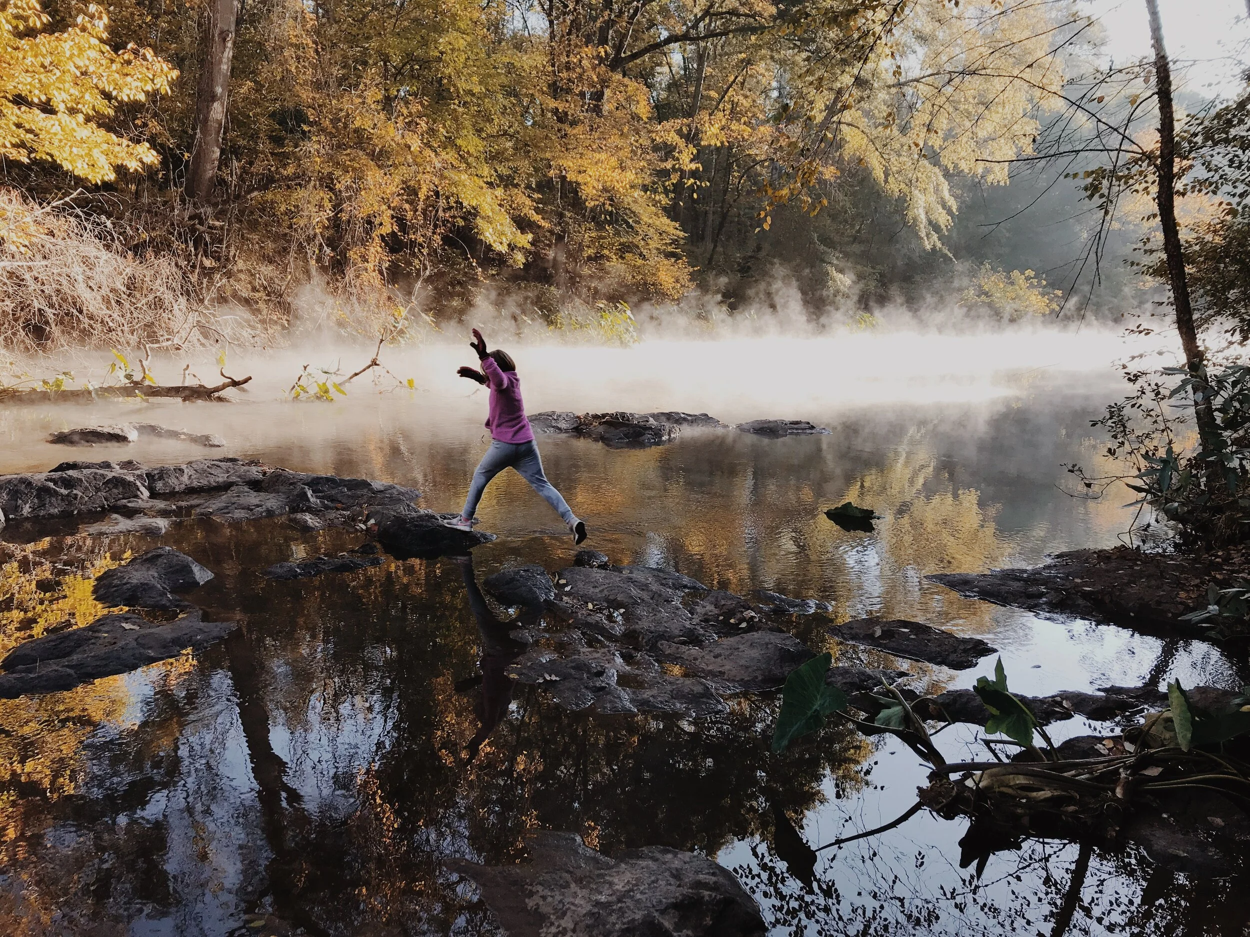 Atlanta family adventure photographer photo of a girl jumping across river rocks on a family hike with fog and sunshine.