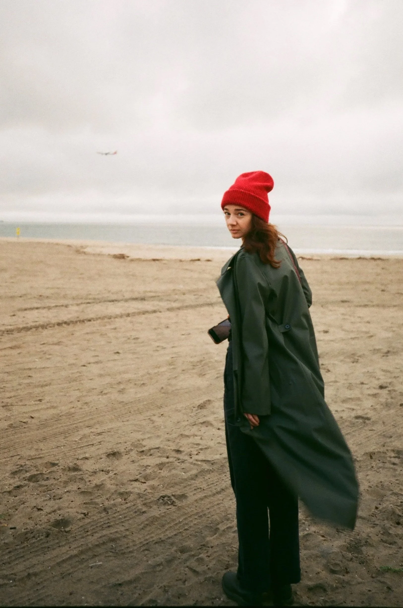 A woman standing on a sandy beach, wearing a red knit hat and a long dark coat, with an ocean and cloudy sky in the background.