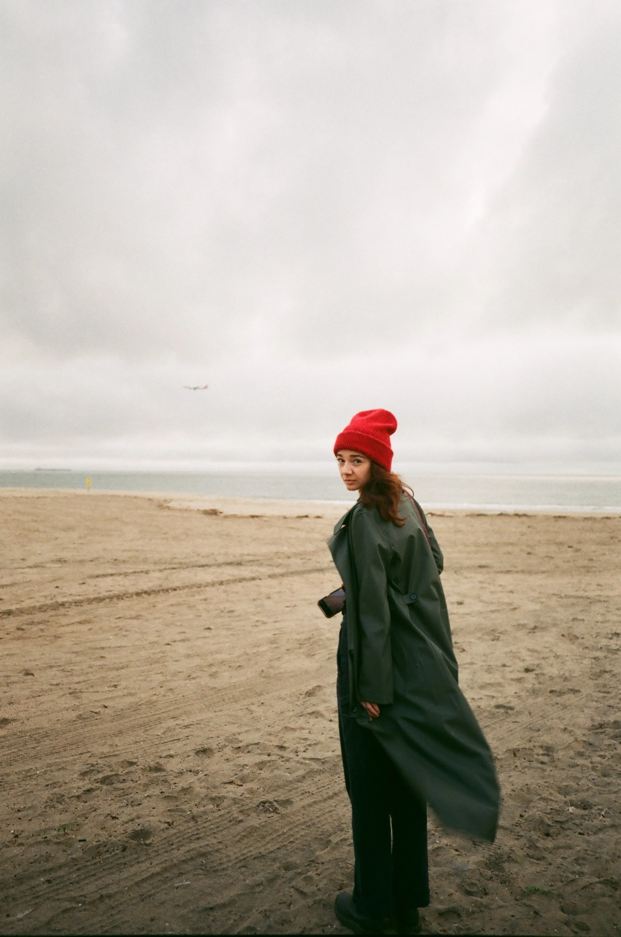 A woman with long brown hair wearing a red beanie, black coat, and black pants standing on a sandy beach on a cloudy day with an airplane flying in the sky in the background.