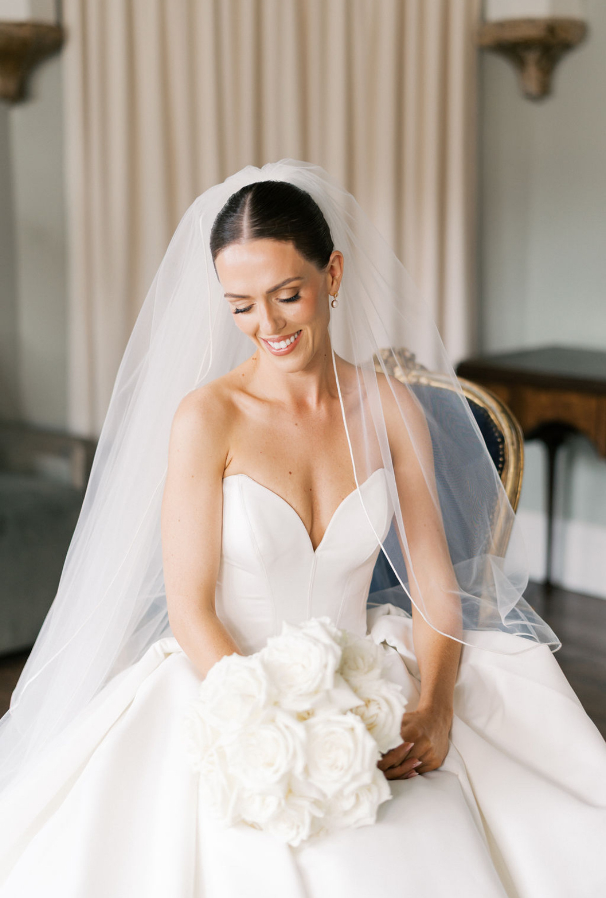 A smiling bride in a strapless white wedding gown with a veil, holding a bouquet of white roses, sitting indoors with cream curtains and vintage furniture in the background.