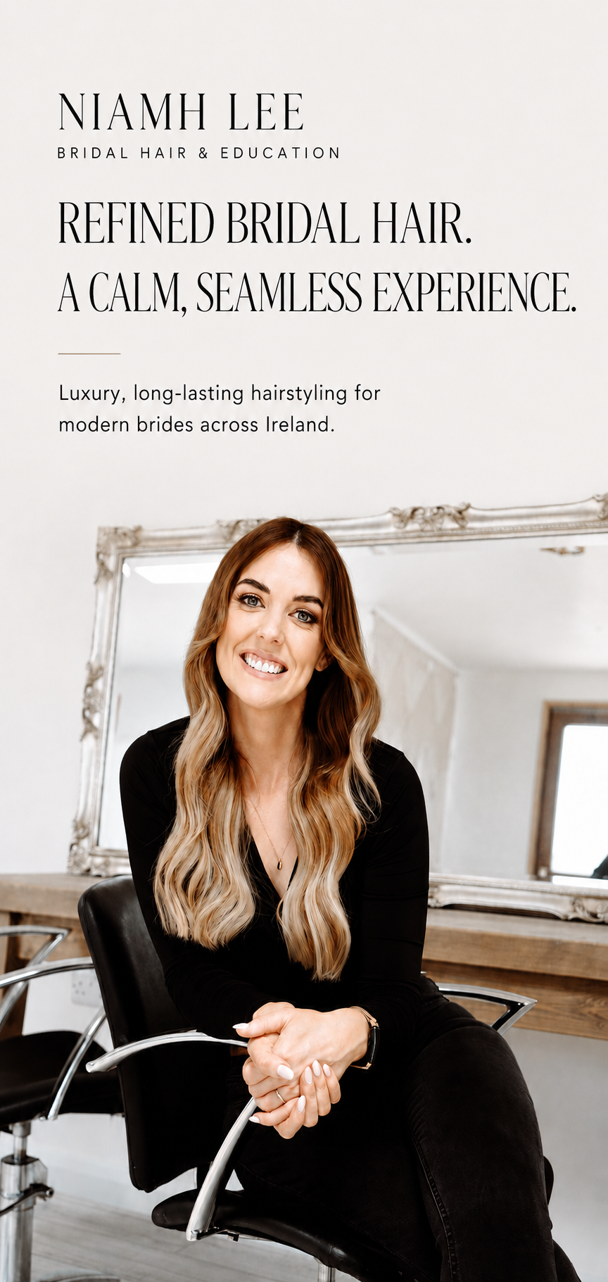 A woman with long wavy blonde hair and a black outfit sitting in a salon chair, smiling. A large mirror is behind her, reflecting a window.