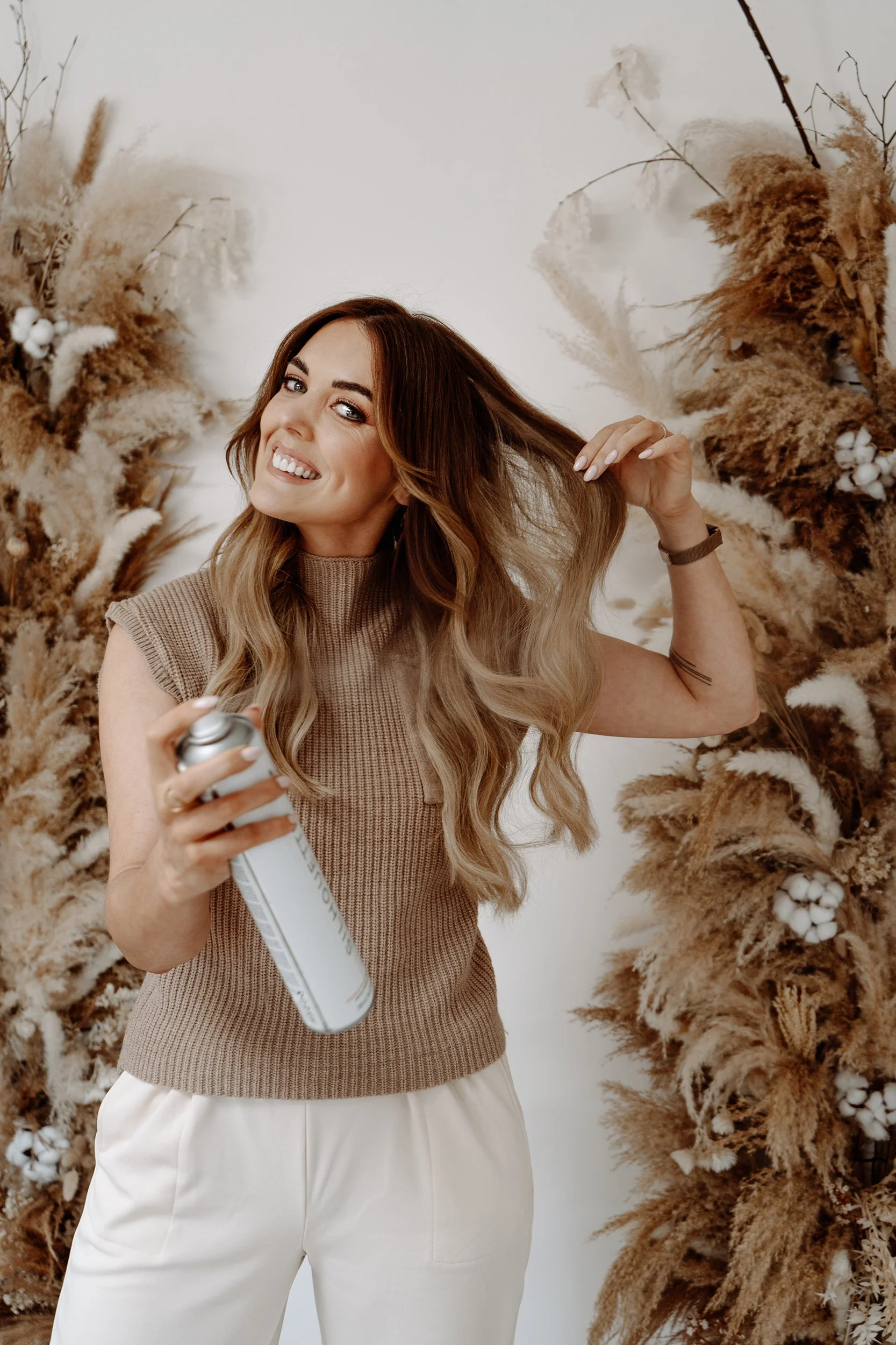 A woman with long wavy hair smiling, holding a can of dry shampoo, and using her hand to touch her hair, standing in front of a beige floral backdrop.