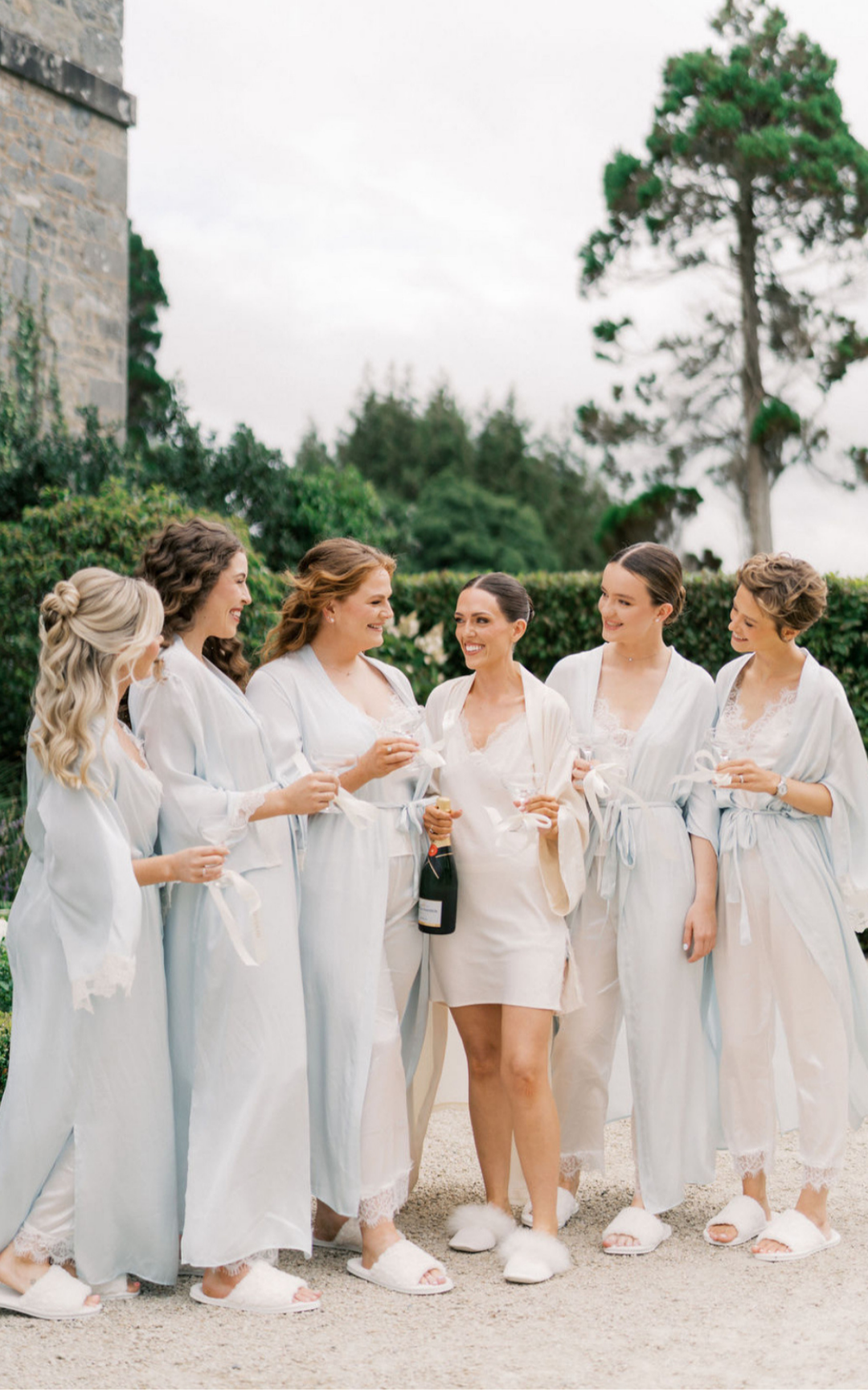 A group of women wearing matching pajamas with lace trim, holding wine glasses, and one holding a bottle, standing outdoors and smiling.
