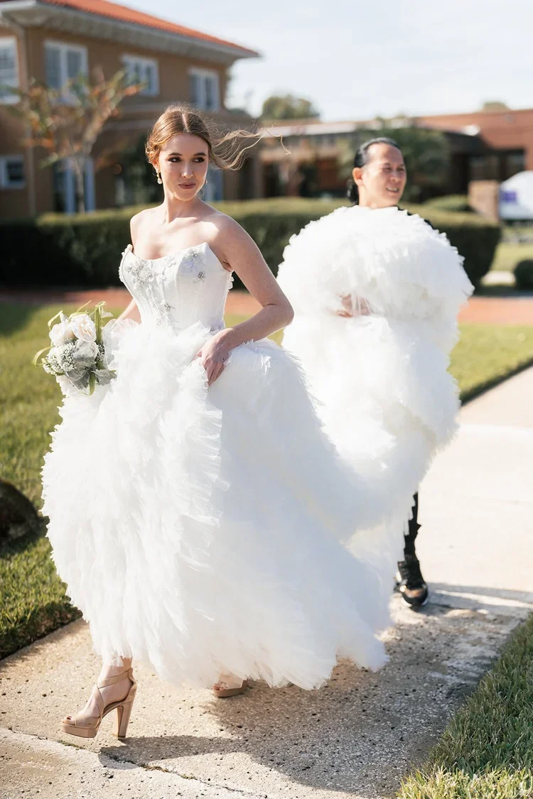Sonthai Gi of Bridal Gown Studio San Jose helping a bride carry the ruffled train of her strapless wedding dress outdoors.