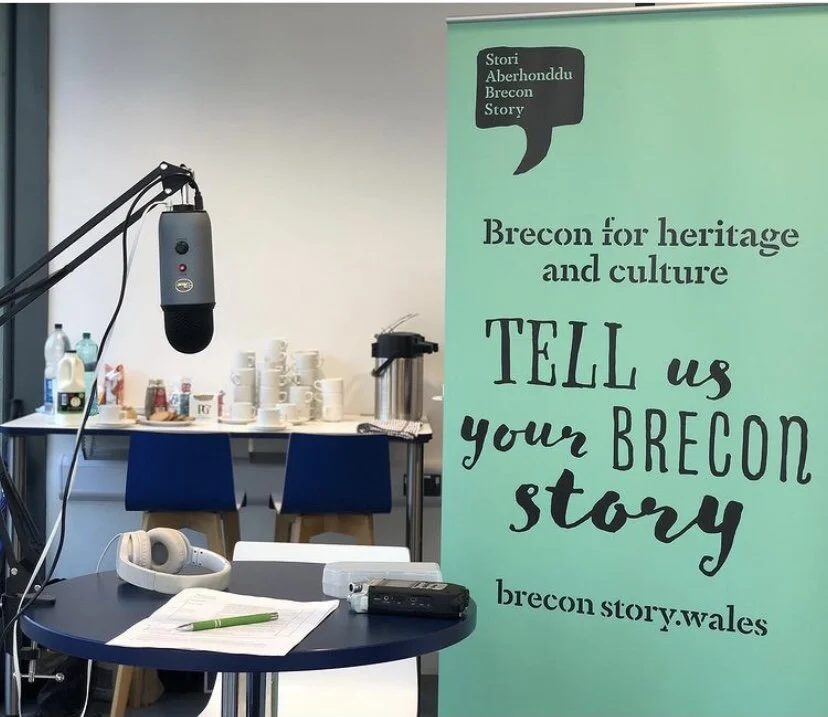 Podcast recording setup with microphone and headphones on a table beside a banner reading “Brecon for heritage and culture — Tell us your Brecon story.”
