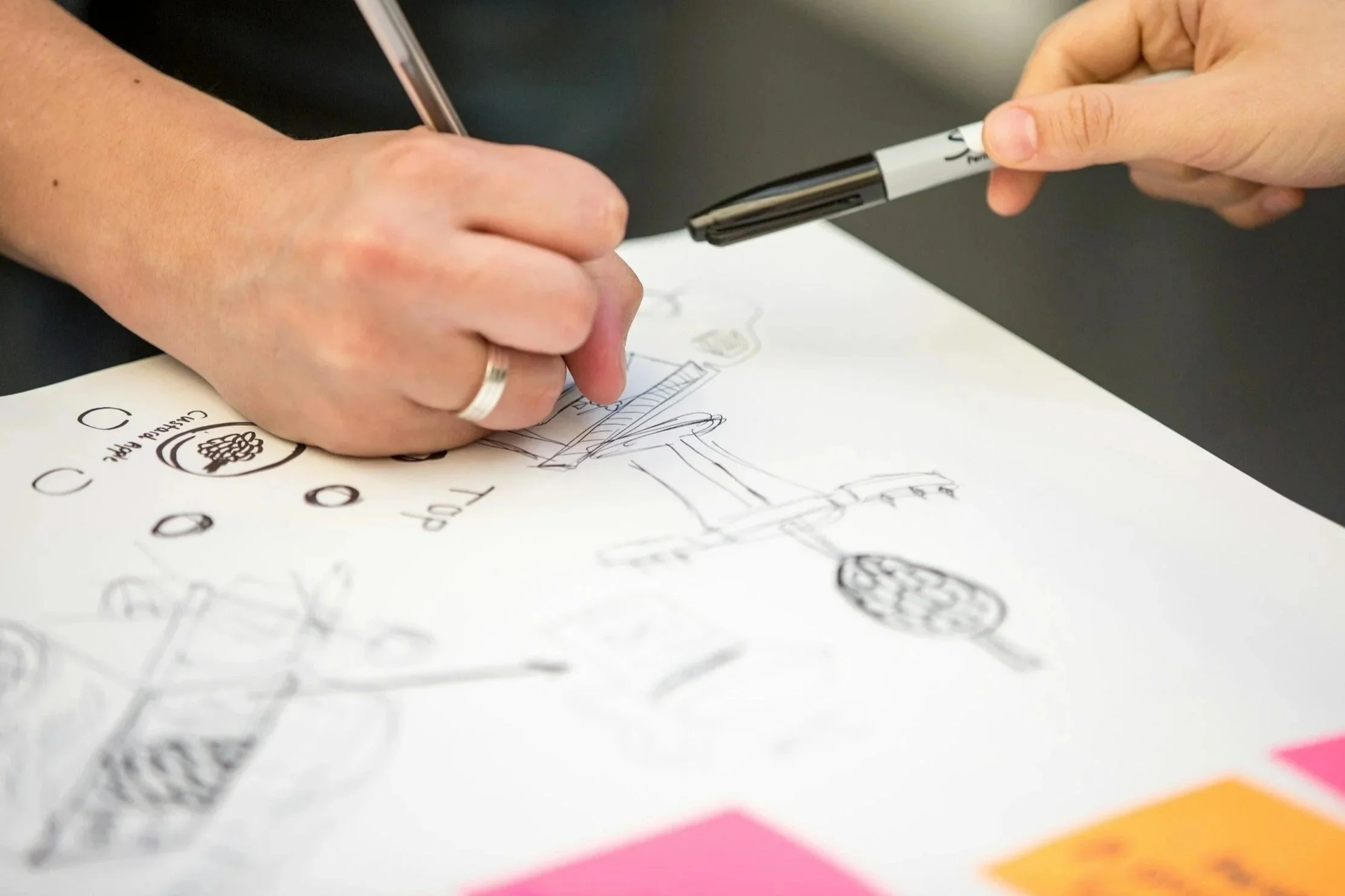 Close-up of a hand sketching interface ideas on paper with pens and markers, showing rough drawings, symbols and notes during a design process.