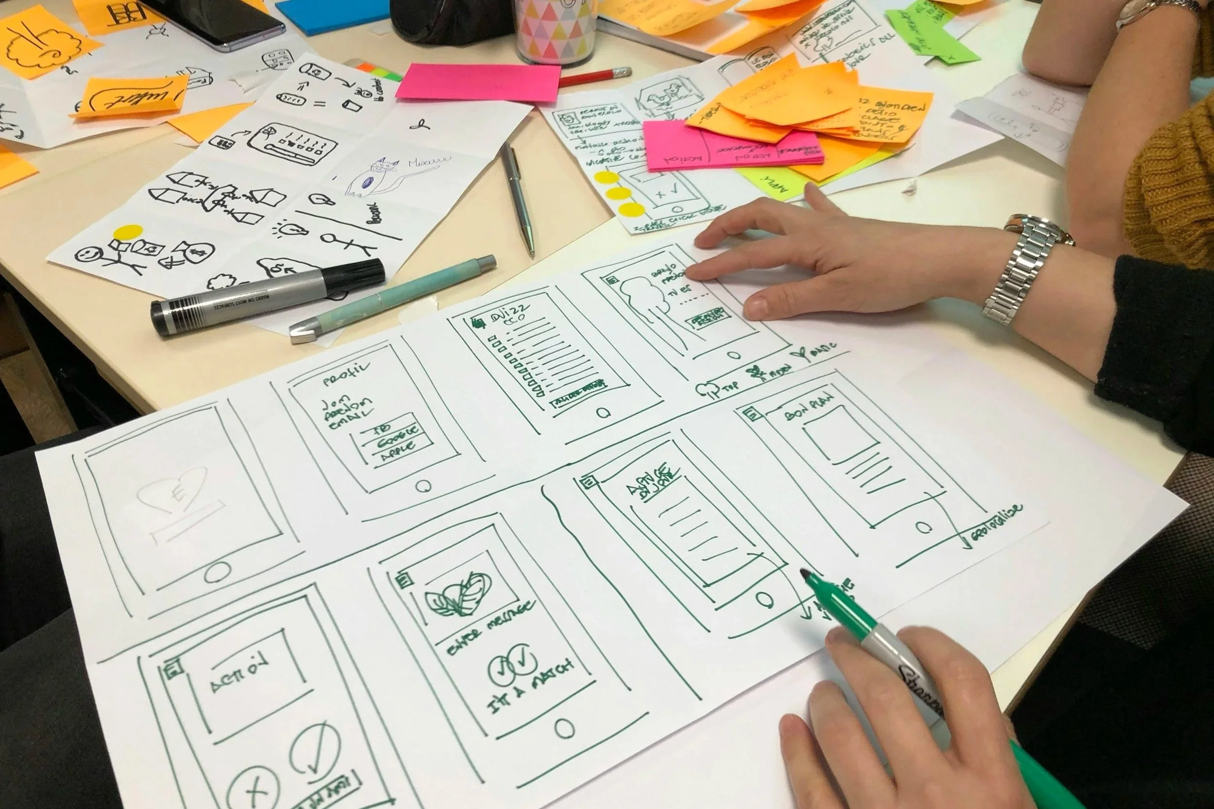 Large sheet of paper spread across a table with hand-drawn mobile app wireframes, surrounded by pens, sketches and sticky notes while participants discuss and annotate the designs.