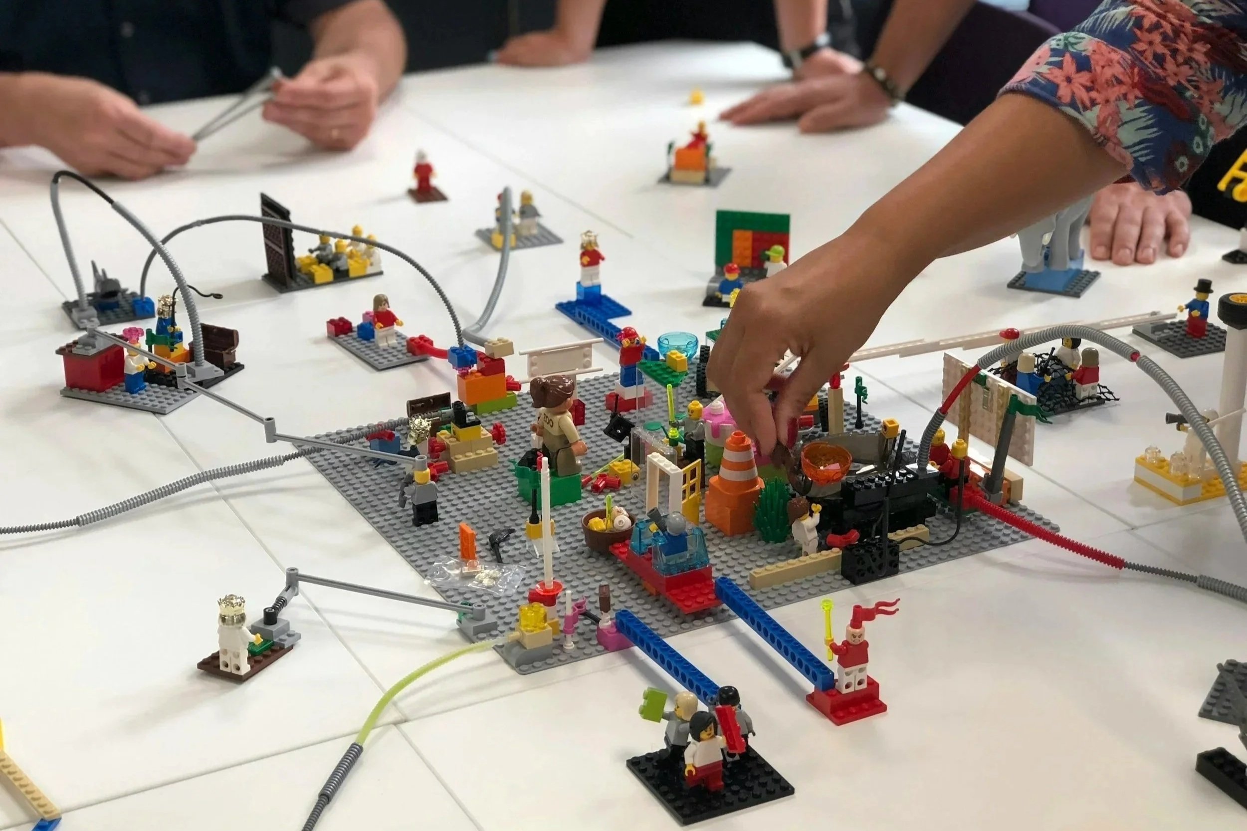 Hands collaborate around a tabletop LEGO scene made of colourful bricks, figures, wires and small objects, as one person adjusts a piece within the miniature environment.