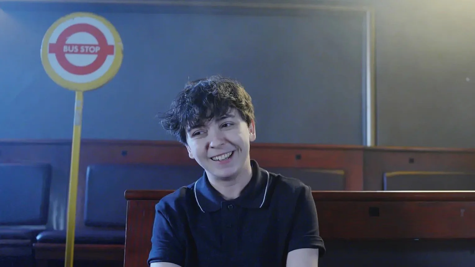 A young person with curly dark hair and a black polo shirt sitting at a bus stop, smiling.