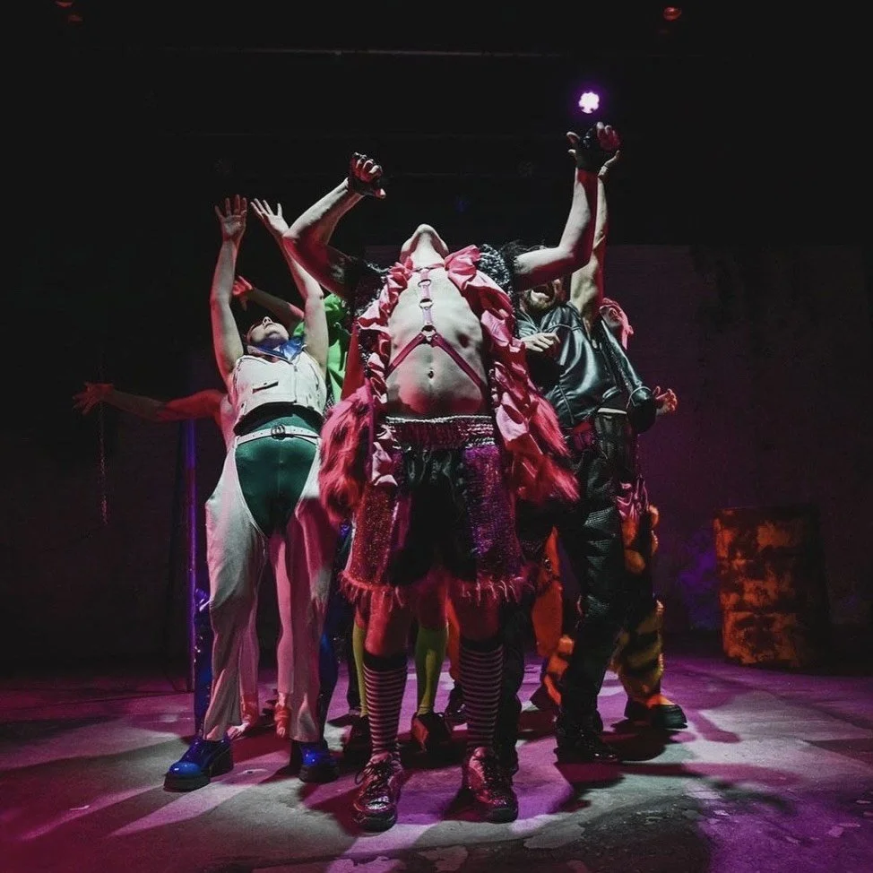 The image shows a group of performers on stage, dressed in vibrant, eclectic costumes with dramatic poses and expressive gestures, under colorful lighting.