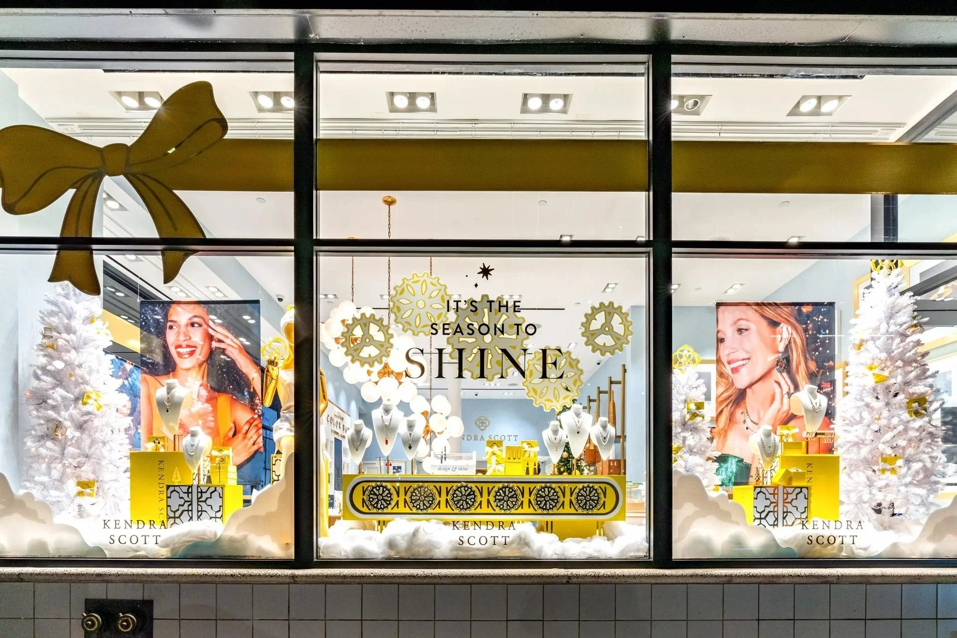 Kendra Scott holiday window display using the brand’s signature colour palette with seasonal greenery and gold accents.