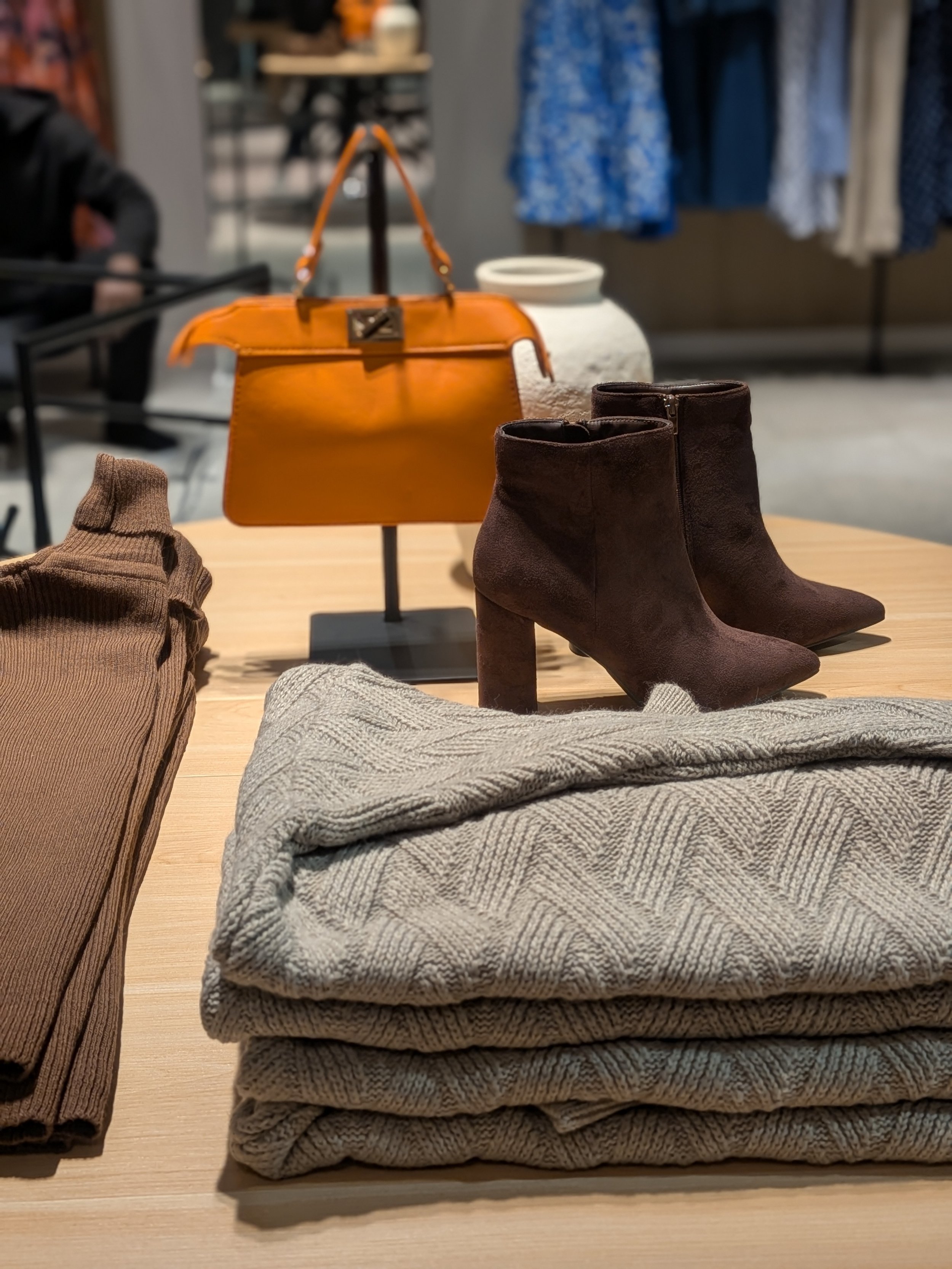 Fashion merchandising display table at Aimee Boutique with handbags, boots, and folded knitwear supporting cross-merchandising and retail flow.