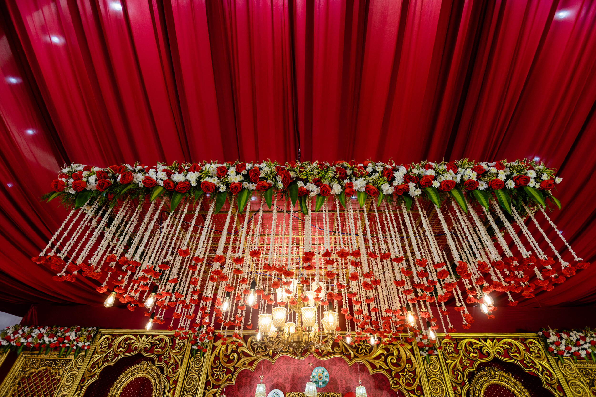 Decorative floral arrangement hanging from a red ceiling at an event, with a chandelier and ornate gold decor below.