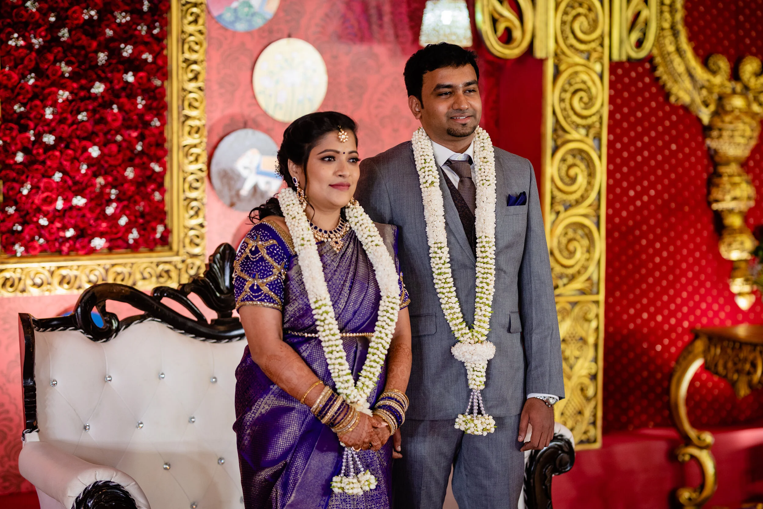 A bride and groom dressed in traditional Indian attire during a wedding ceremony, standing together adorned with floral garlands, with ornate red and gold decorations in the background.