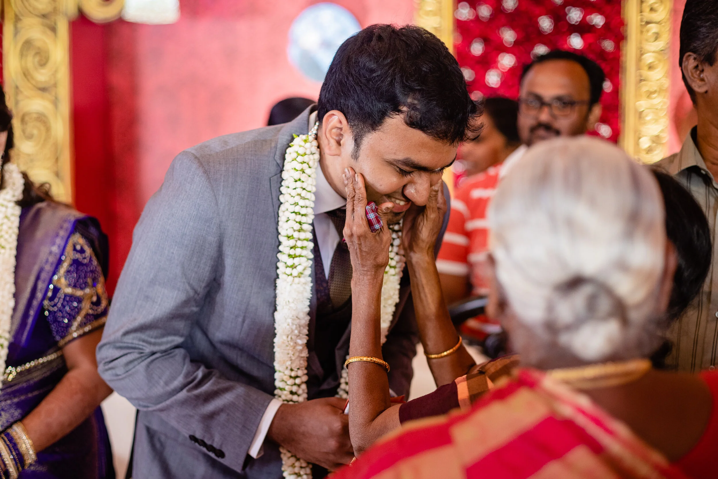 A man and an elderly woman touching faces during a traditional celebration with colorful background and garlands, in a festive gathering.