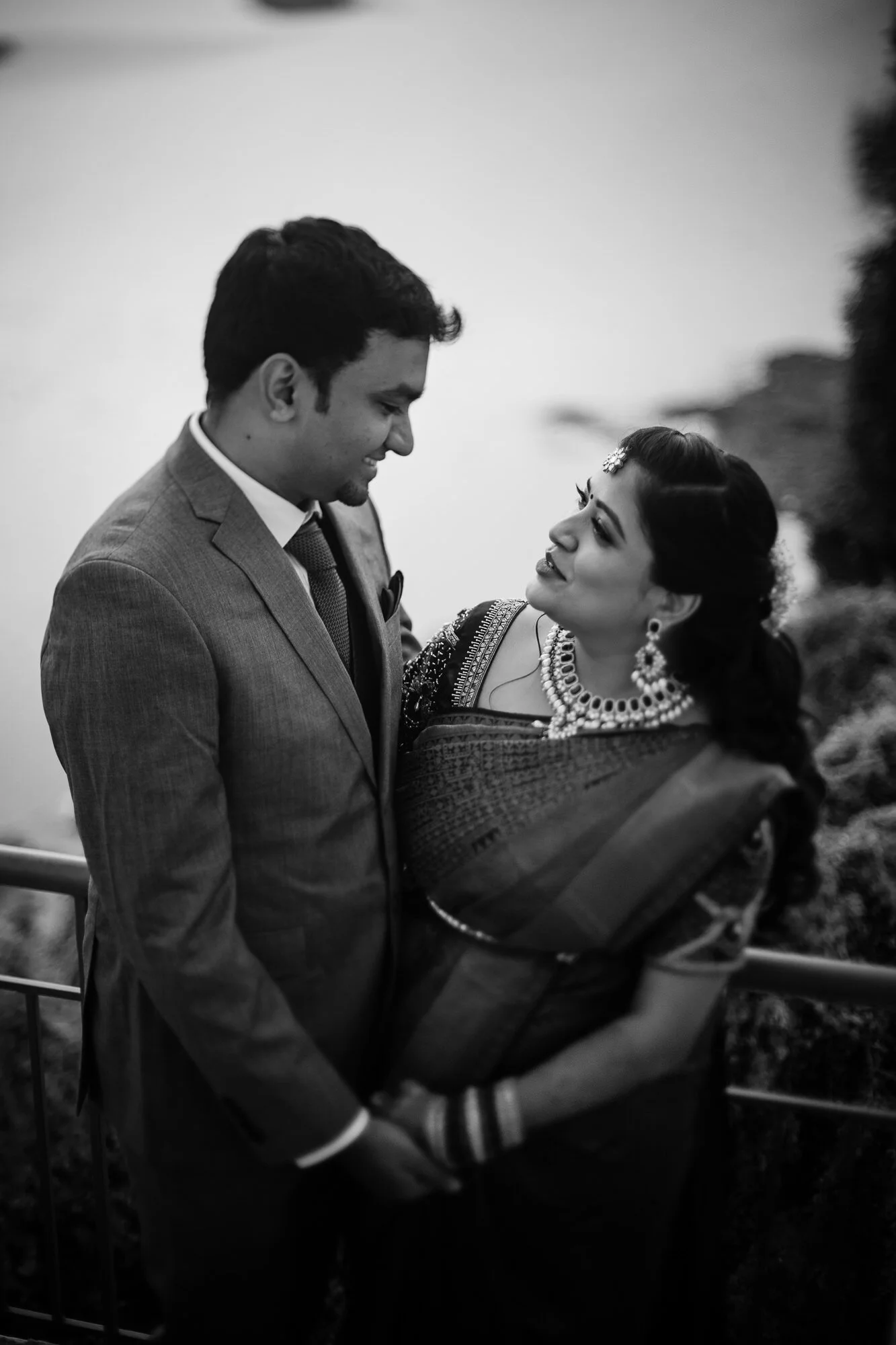 A black and white photo of a man in a suit and a woman in traditional Indian attire looking at each other affectionately, standing on a balcony with some trees in the background.