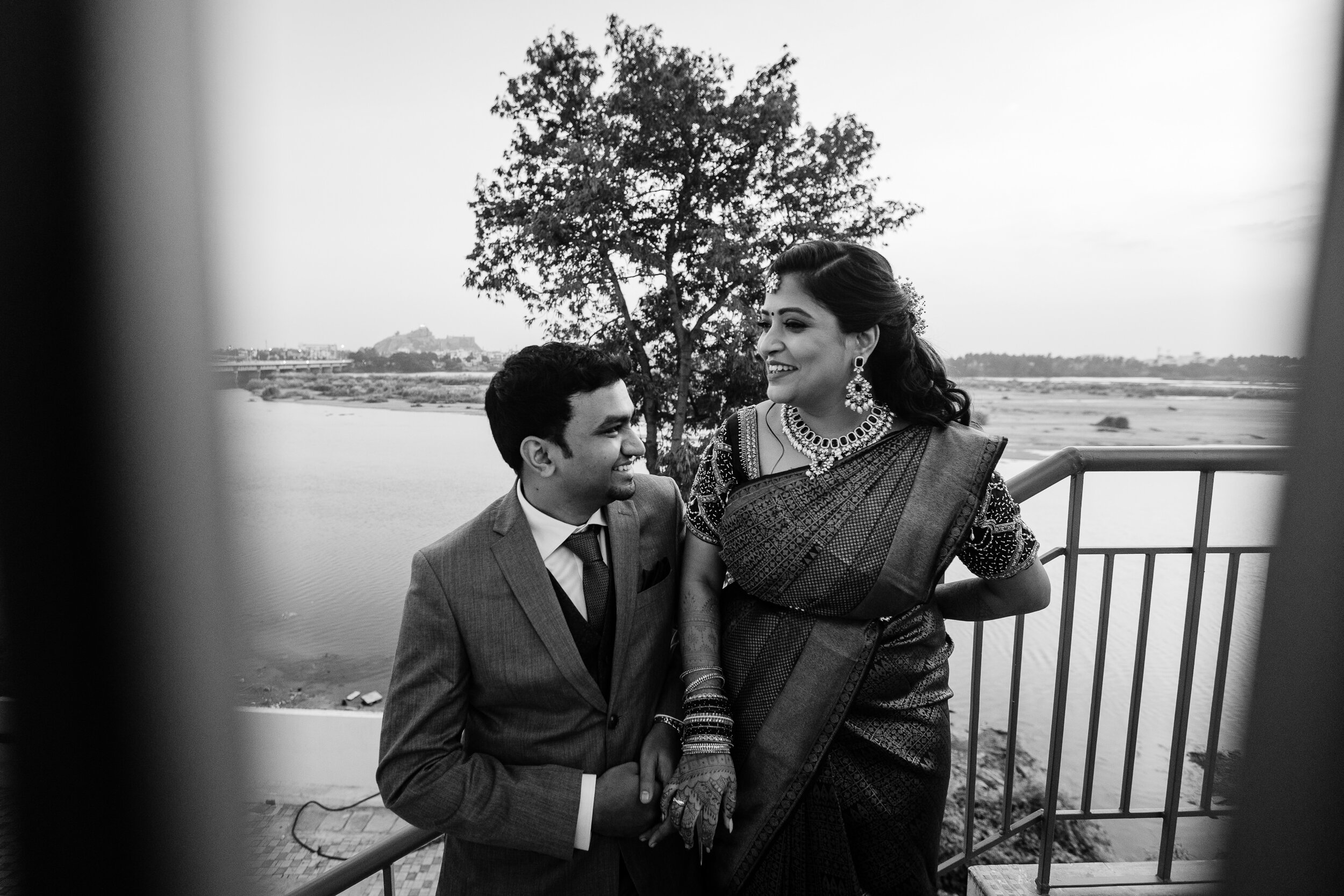 A black and white photo of a man and woman smiling, dressed in traditional Indian attire, standing on a balcony with a lake and trees in the background.
