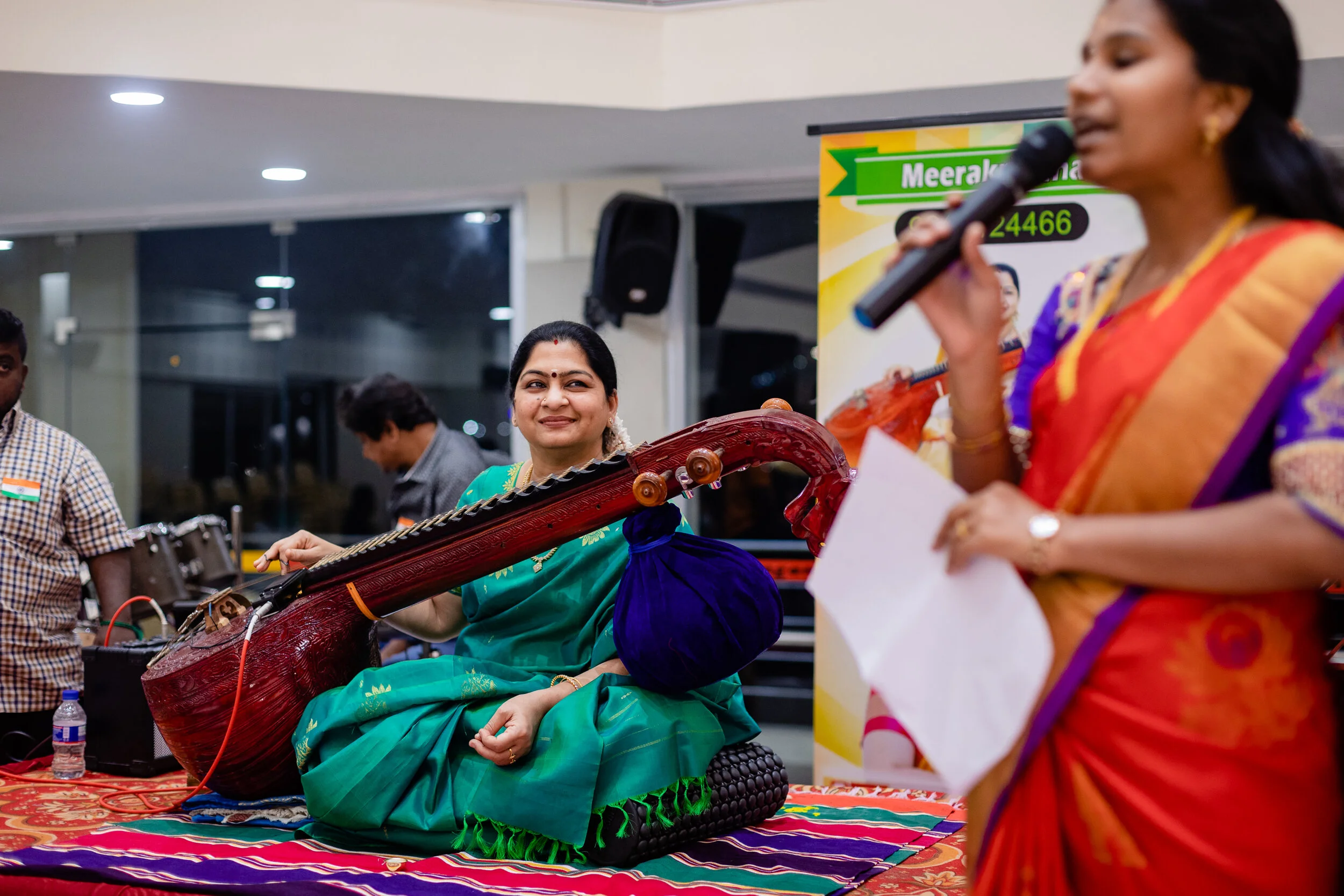 A woman in traditional Indian attire playing a sitar at an event, with another woman speaking into a microphone in the foreground.