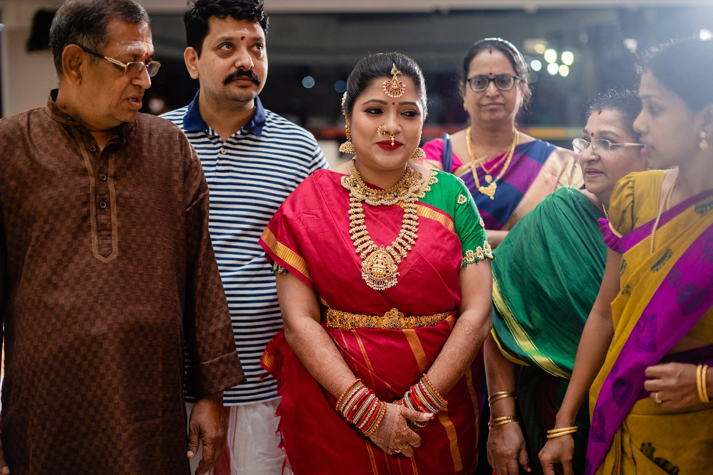 A group of women and men dressed in traditional Indian sarees and kurta at a cultural or religious event. The woman at the center wears a red saree with gold jewelry and henna on her hands, surrounded by others also dressed in colorful sarees and tra