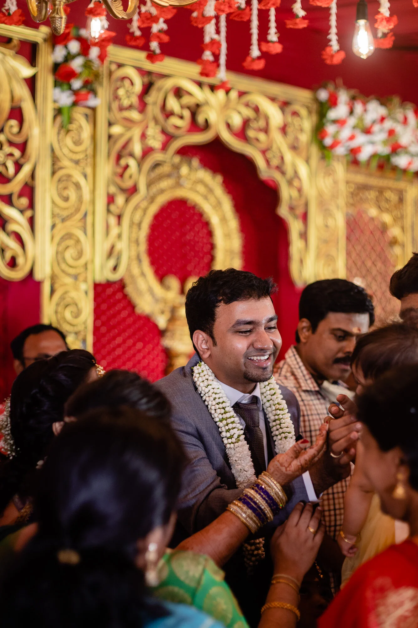 A group of people participating in a traditional Indian wedding ceremony, with the groom in a suit and wearing a flower garland, surrounded by women in colorful sarees and jewelry, in a decorated wedding venue with red and gold accents.