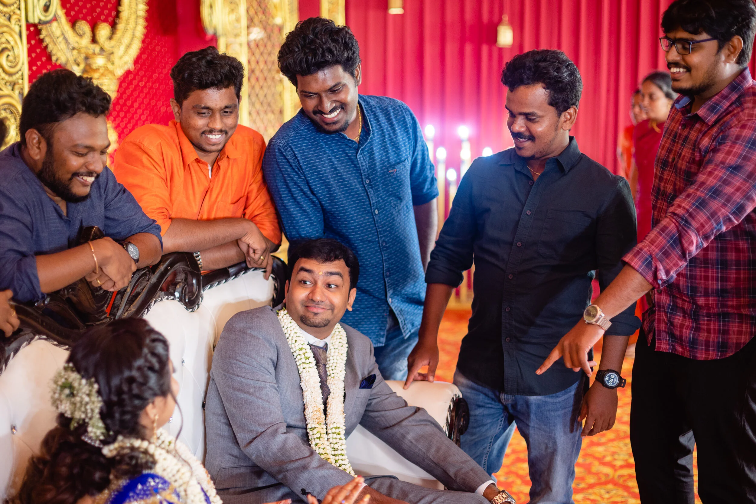 Group of men and women gathered around a seated groom at a wedding celebration, with red and gold decorated background.
