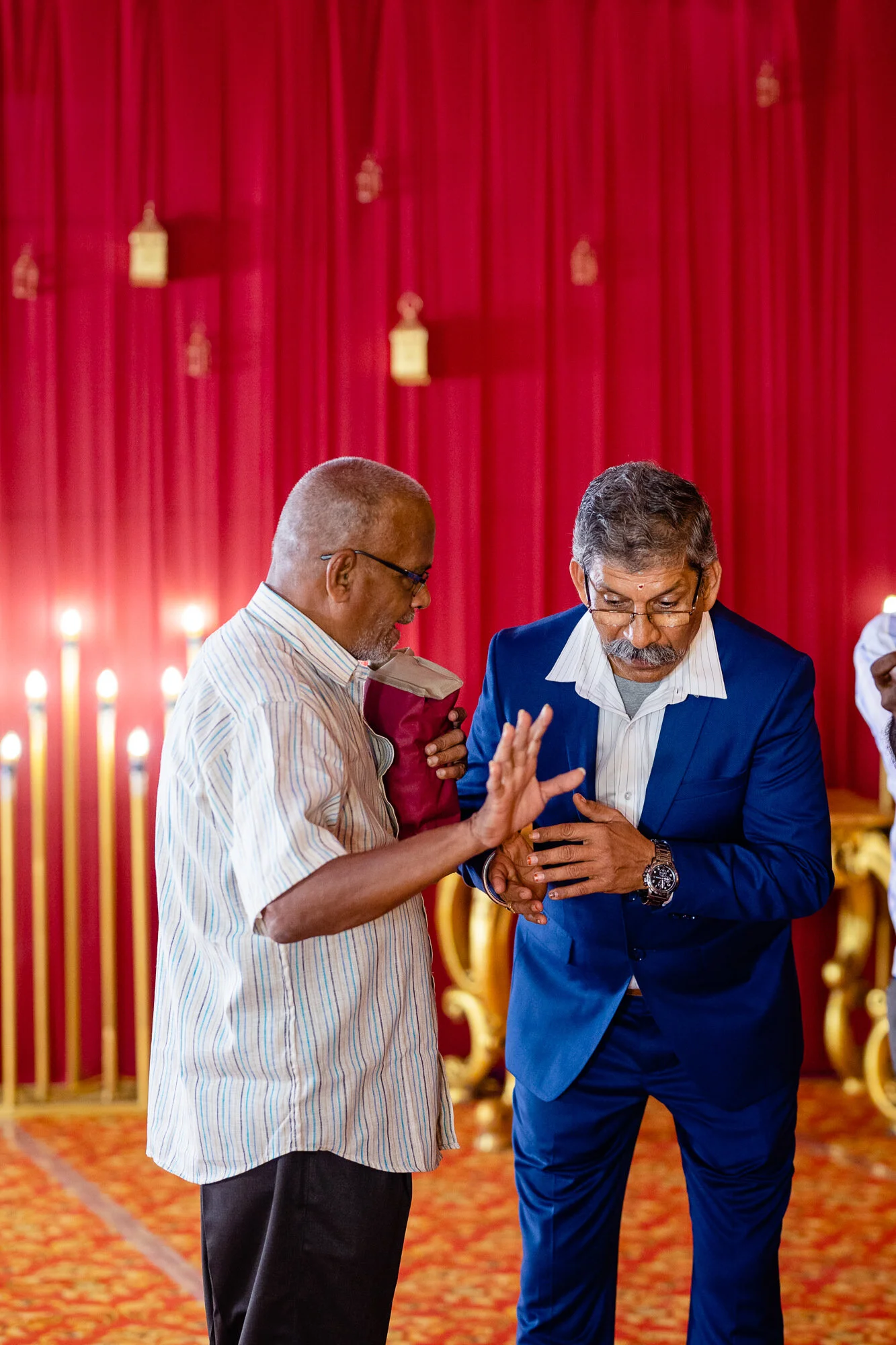 Two men dressed in formal attire engaged in conversation at a festive event with red curtains and yellow decorations in the background.