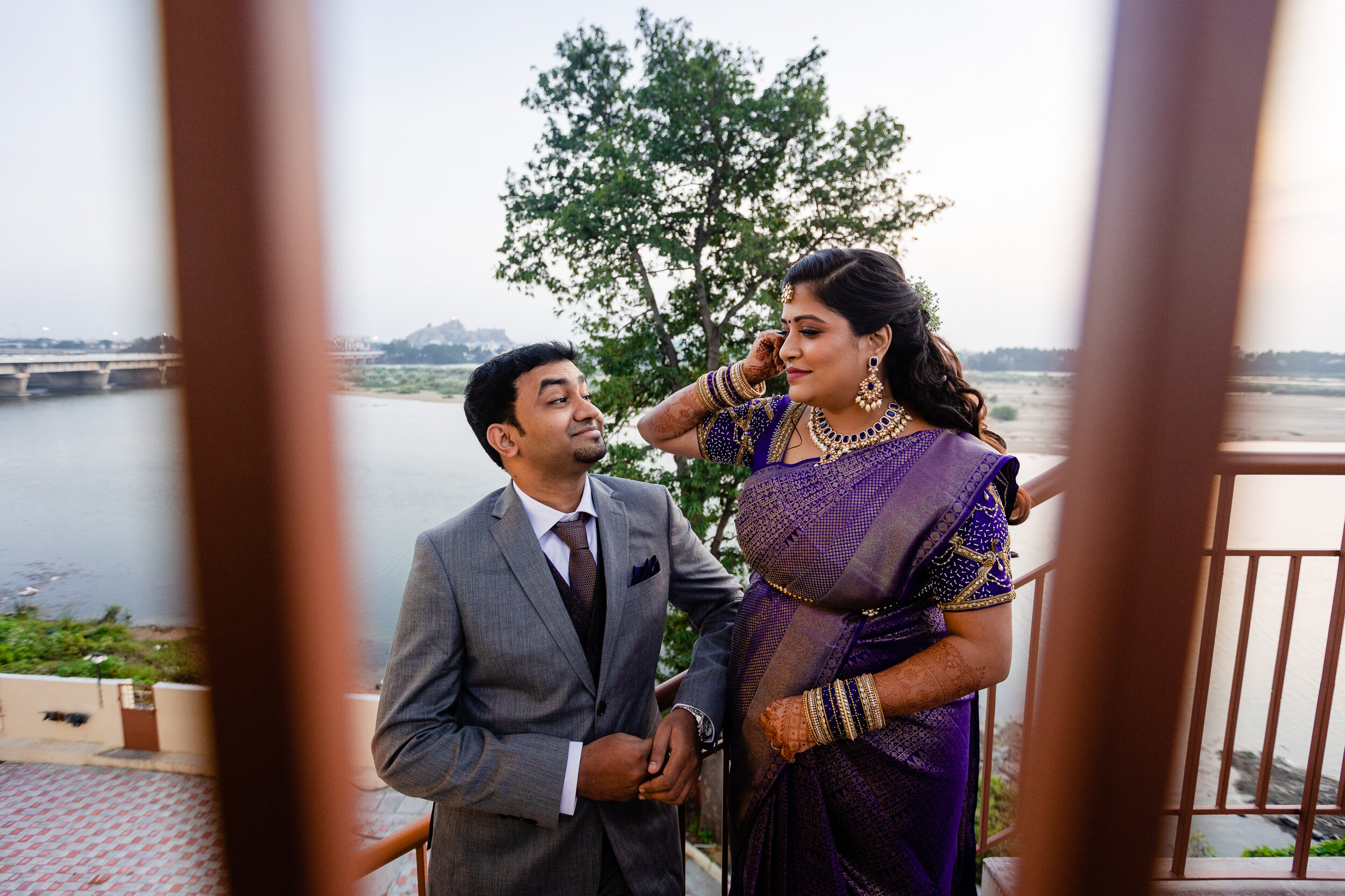 A man in a gray suit and a woman in a purple saree with gold jewelry standing on a balcony near a river, with trees and a distant bridge in the background.