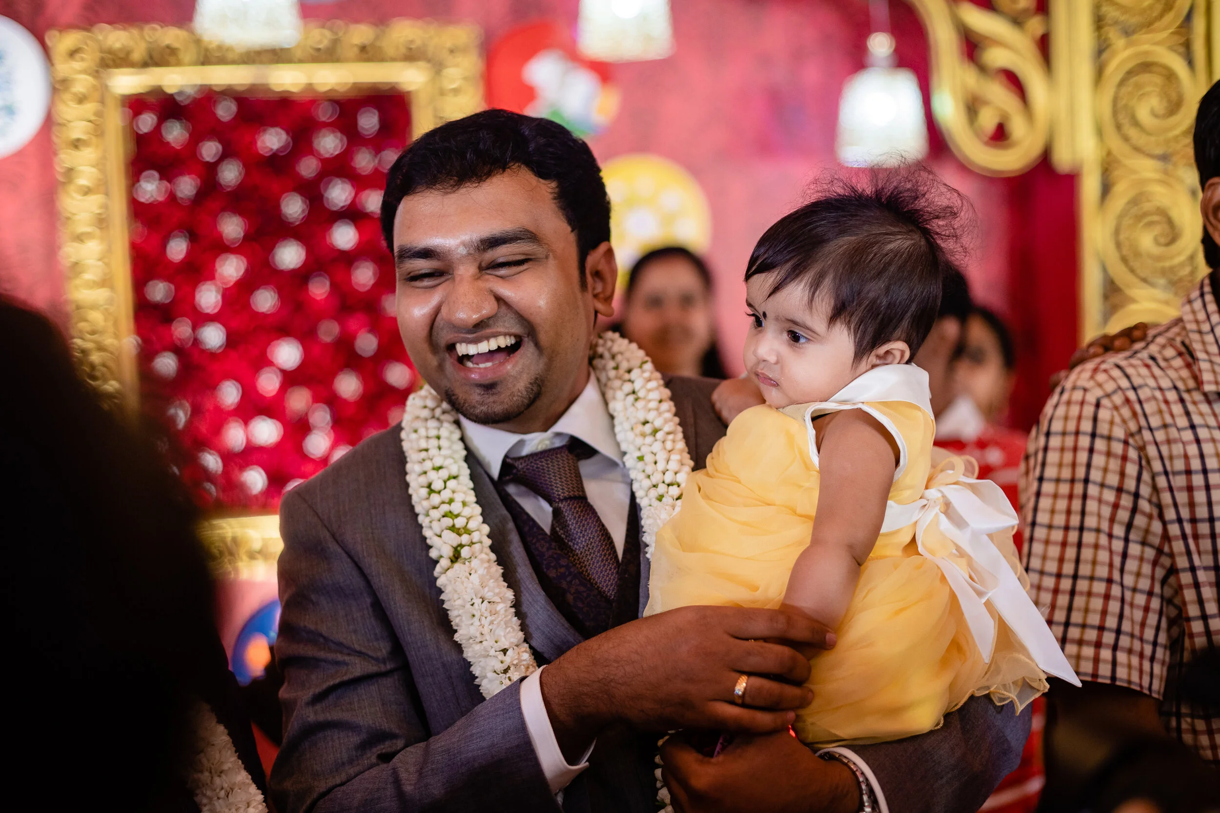 A man in a suit holding a young girl in a yellow dress at a festive celebration, with a decorated red backdrop and other guests in the background.