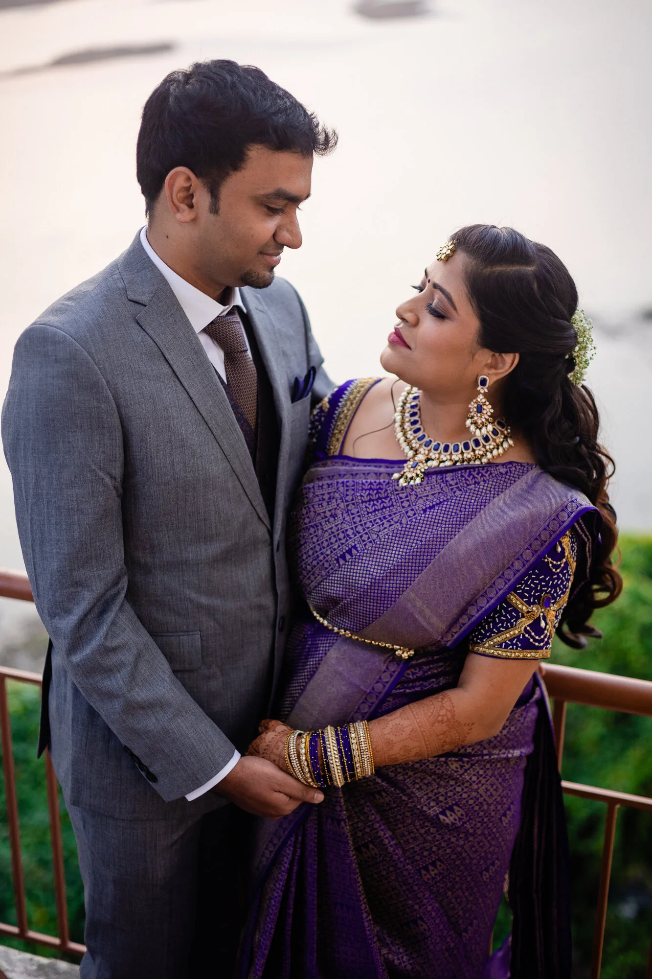 A couple dressed in formal wear, with the woman in traditional Indian attire and jewelry, holding hands and standing close to each other by a railing near water.