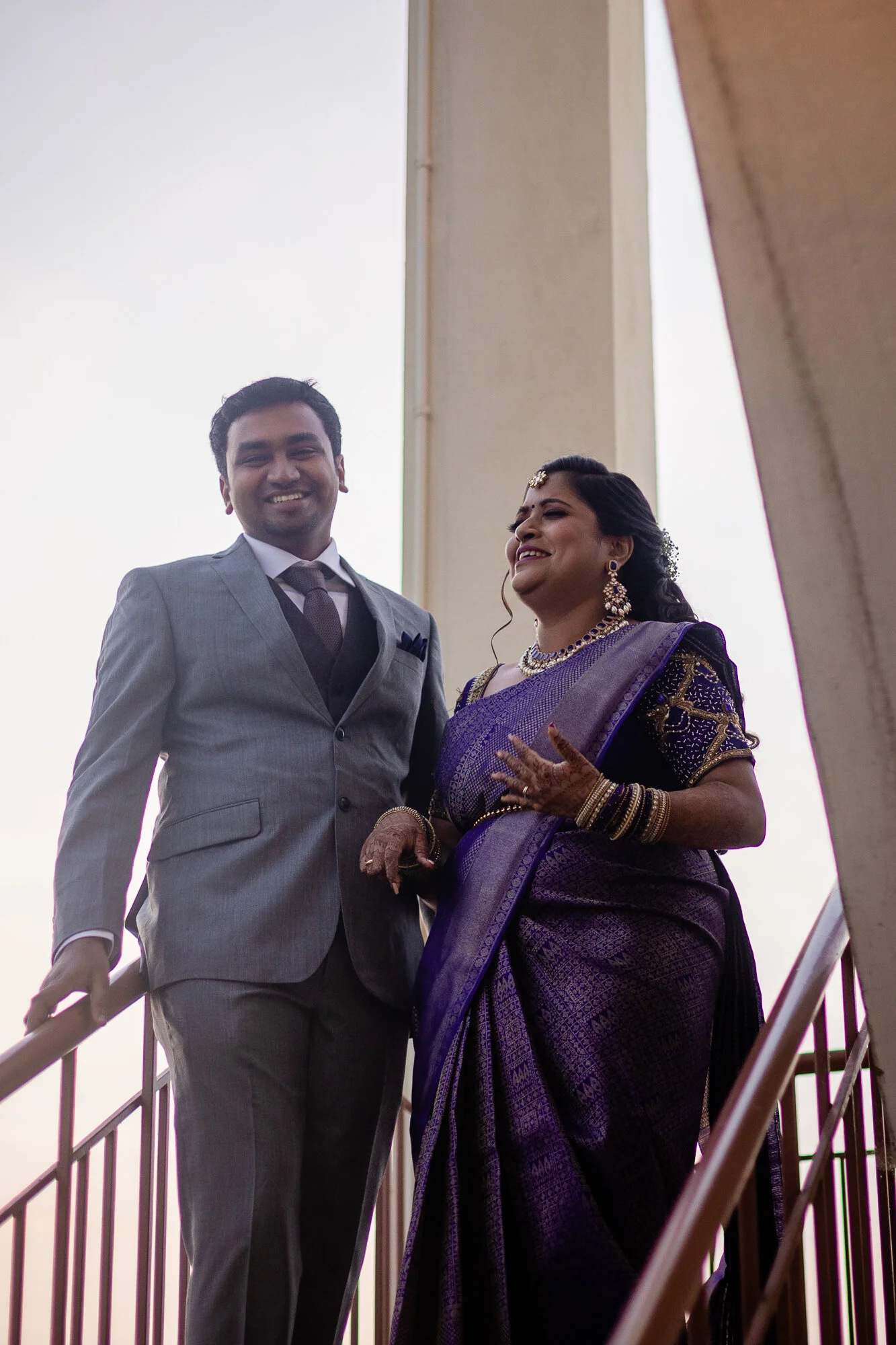A man and woman wearing traditional and formal attire standing on a staircase, smiling and looking happy.