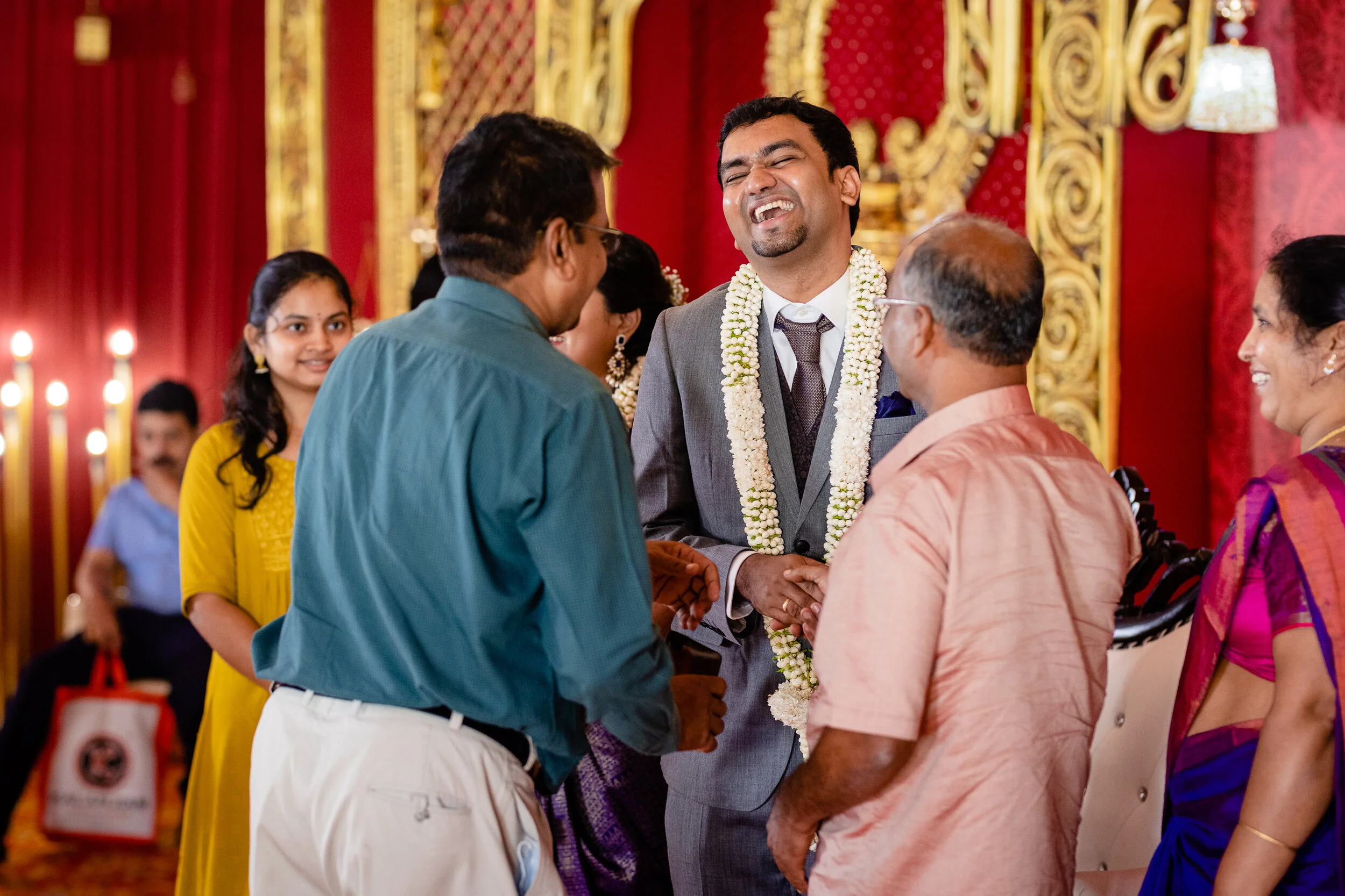A groom in a gray suit with a white garland around his neck, smiling and holding hands with a man, at an Indo-Aryan wedding ceremony, surrounded by family members in colorful traditional clothing, in an ornate decorated wedding hall with red and gold
