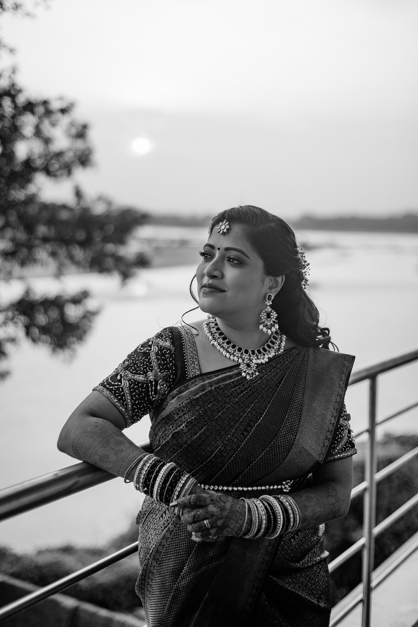 A woman dressed in traditional Indian attire, wearing jewelry and bangles, standing outdoors on a balcony or terrace overlooking a river or lake during sunset in black and white.