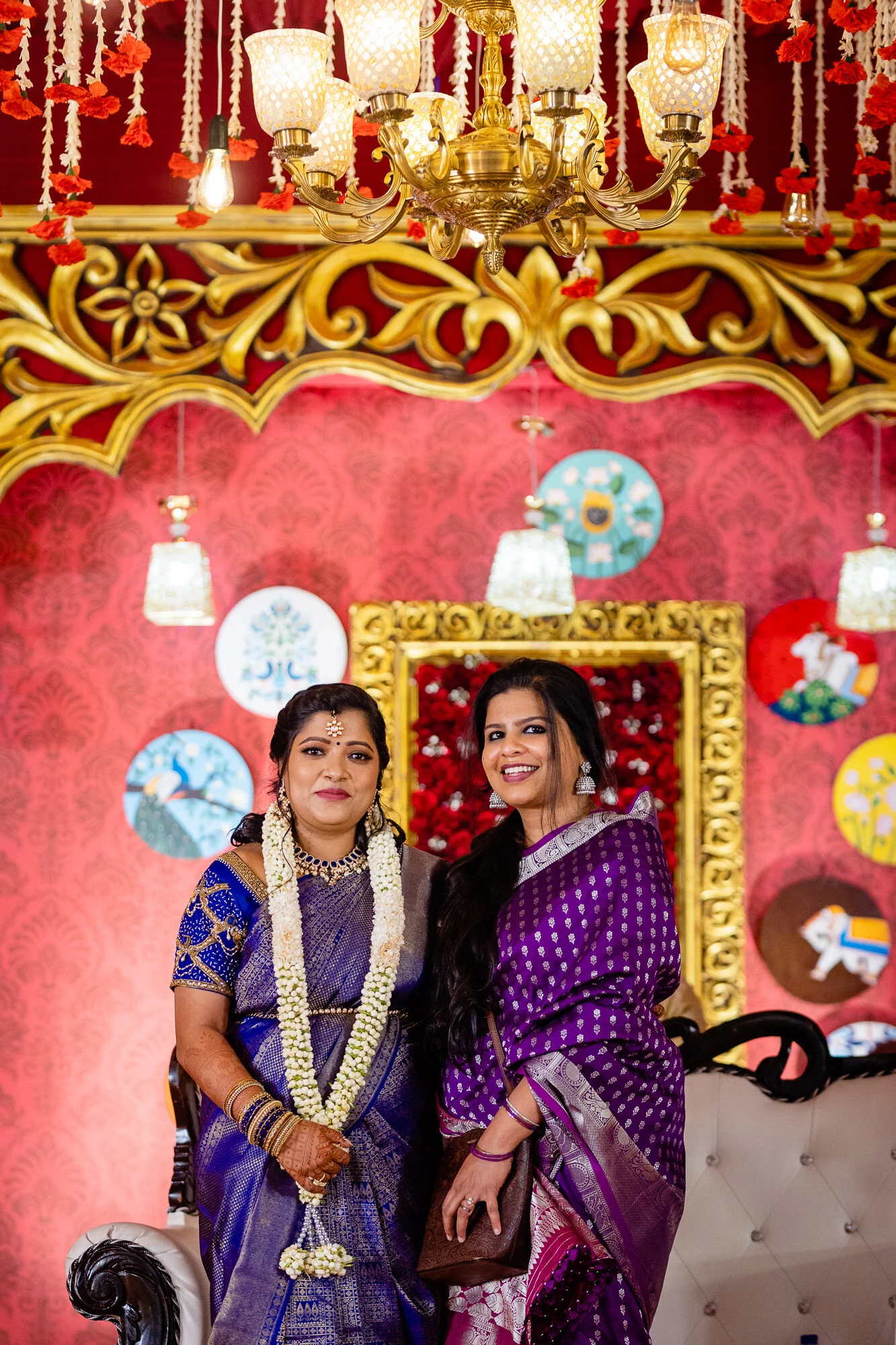 Two women wearing traditional Indian sarees posing at a decorated event with red and gold background, floral and elephant motifs, and ornate lighting fixtures.