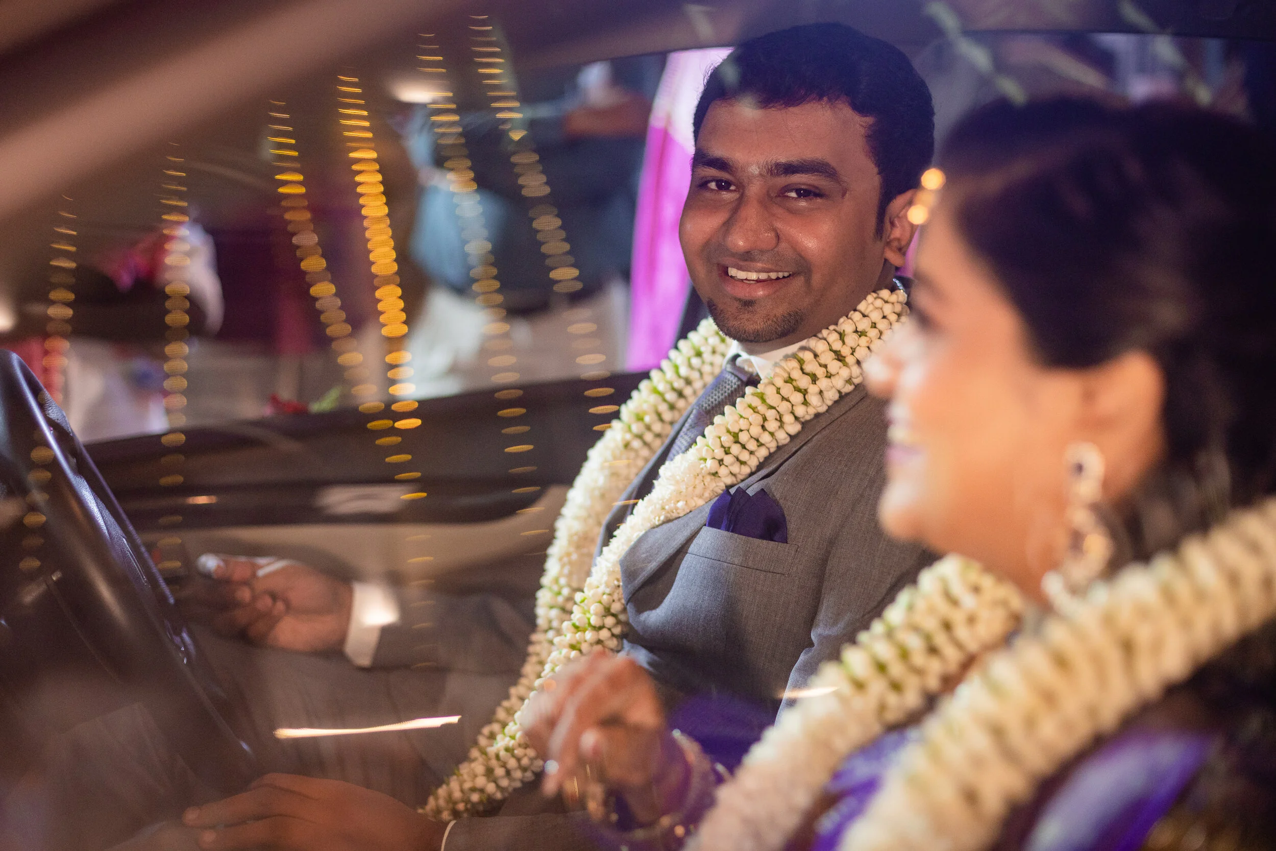 A groom and bride sitting inside a car, both wearing flower garlands, smiling and enjoying a wedding celebration, with blurred festive lights in the background.