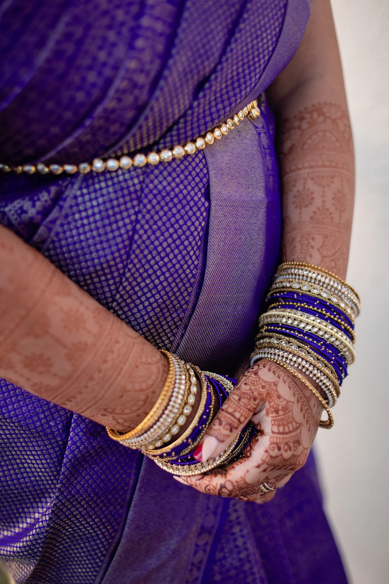 Close-up of a woman's hands and waist, adorned with traditional gold and pearl jewelry, wearing a purple saree with detailed henna designs on her arms.