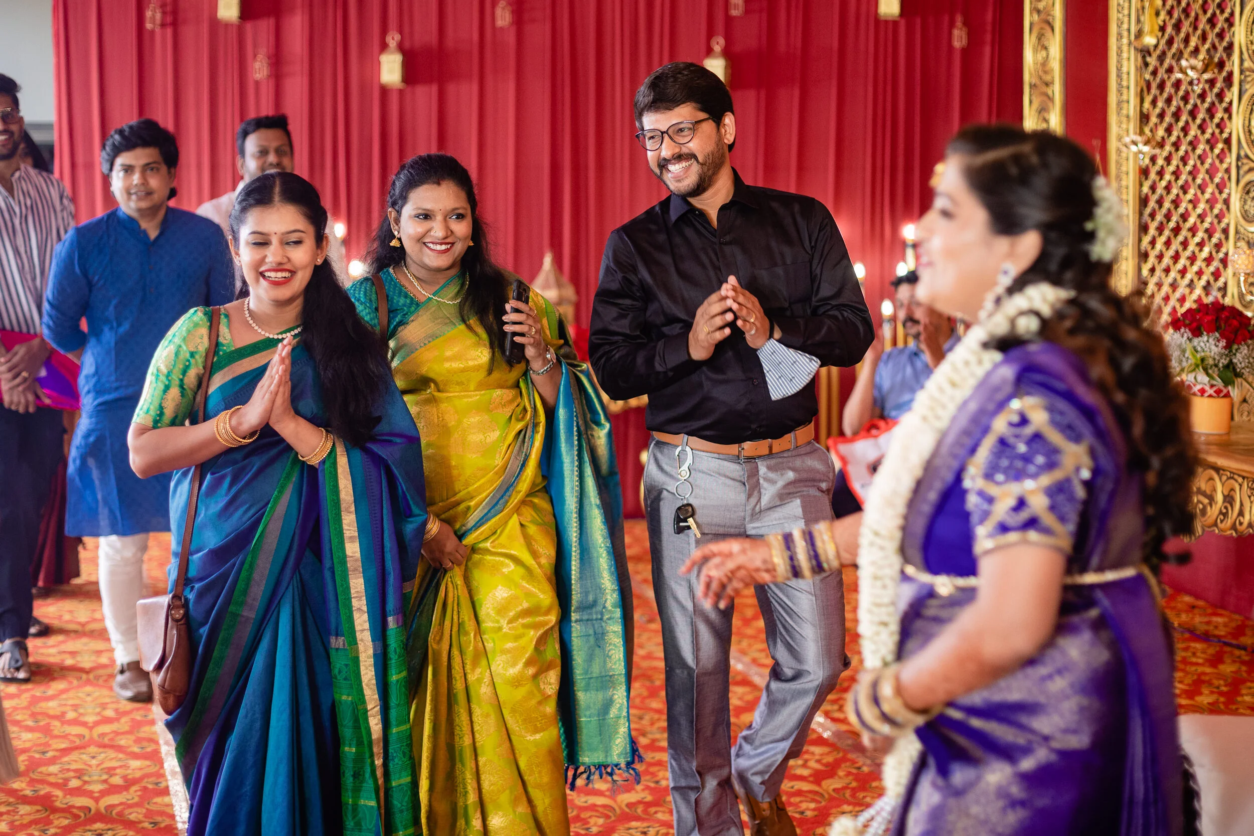 Group of people at a celebration or wedding, dressed in traditional colorful Indian attire, greeting and smiling.