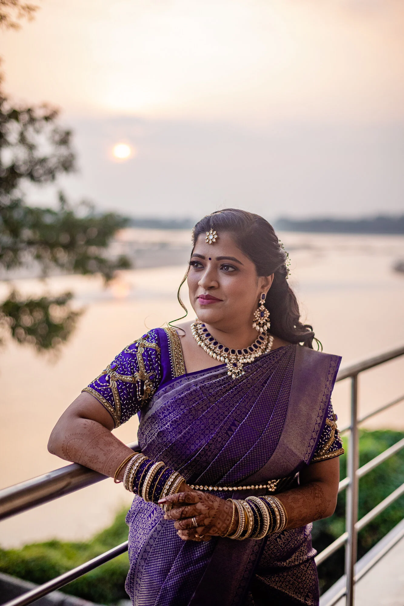 A woman dressed in a traditional purple saree and elaborate jewelry poses outdoors near a river at sunset.