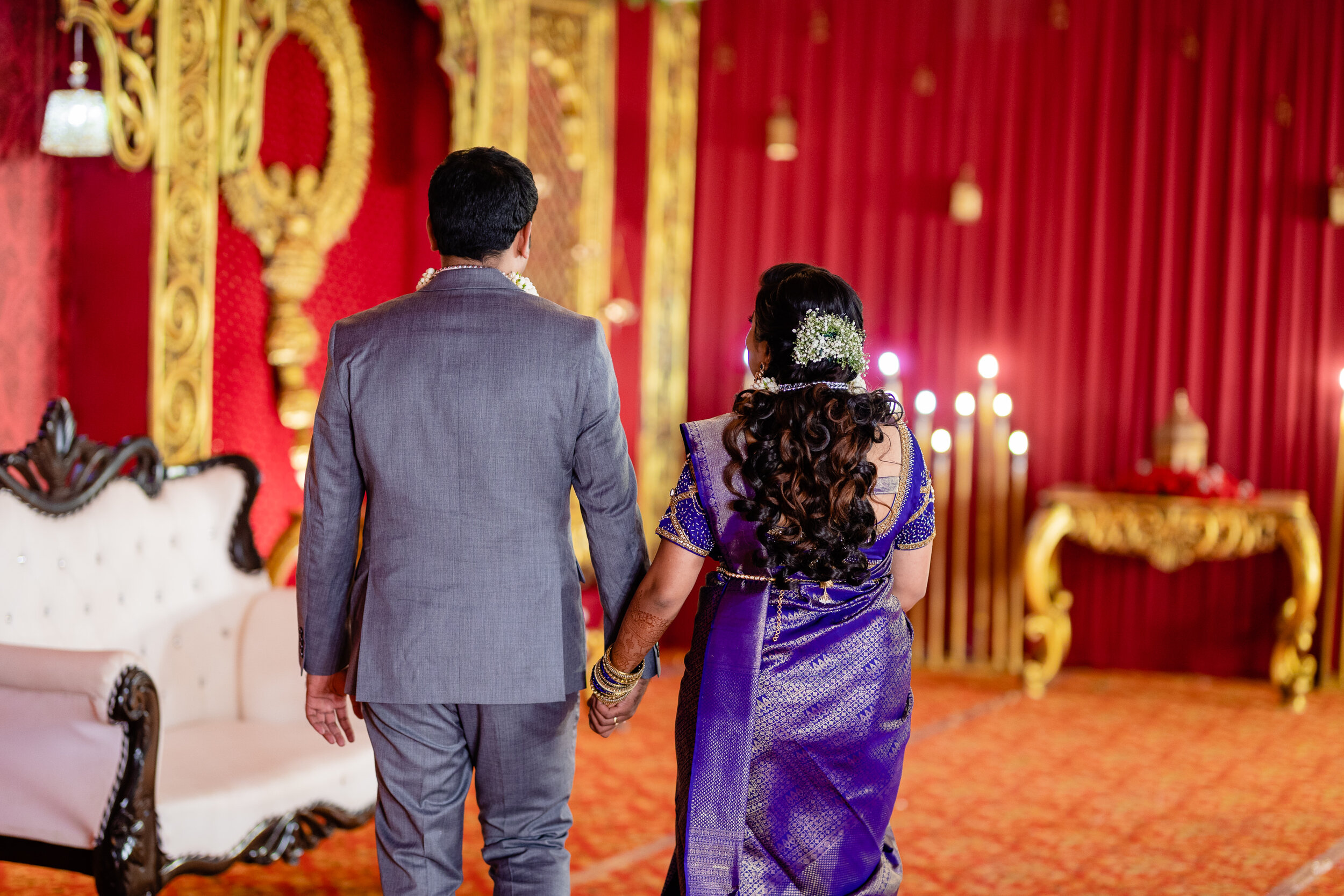 A couple holding hands in a luxurious red and gold decorated room, possibly at a wedding or traditional celebration.